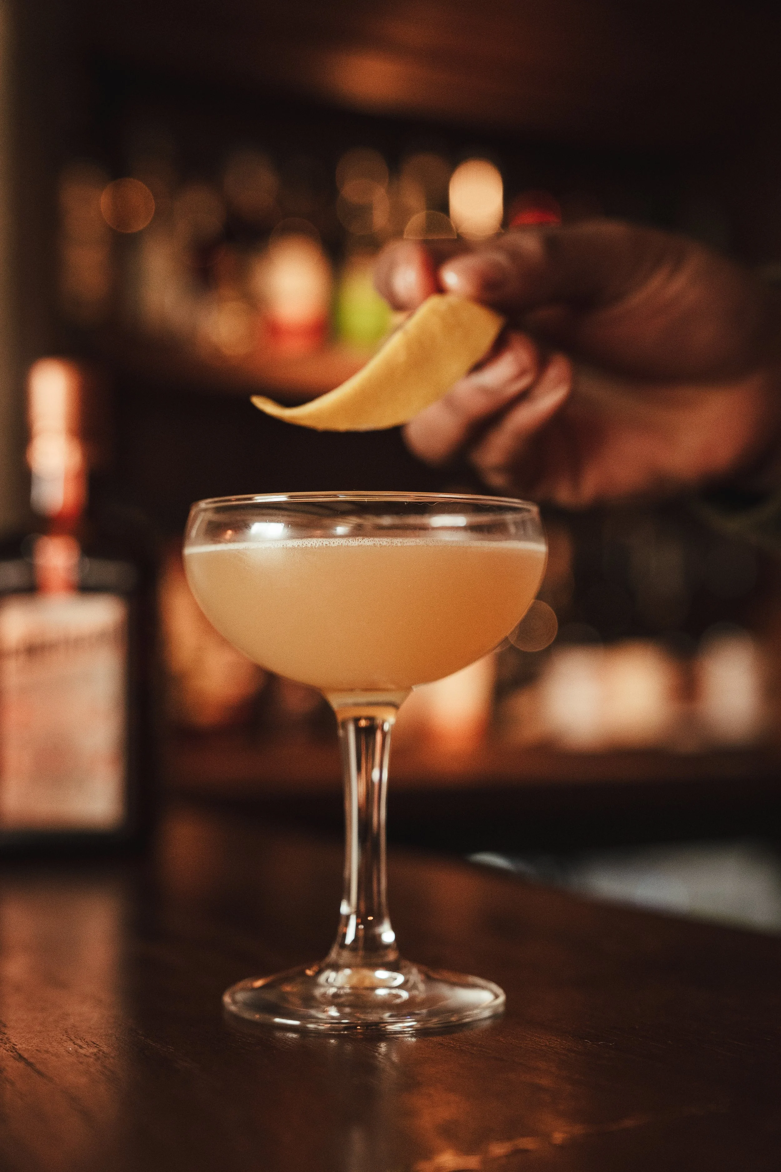 Close-up of a cocktail in a coupe glass with a slice of lemon being added by a bartender with a blurred bar in the background.