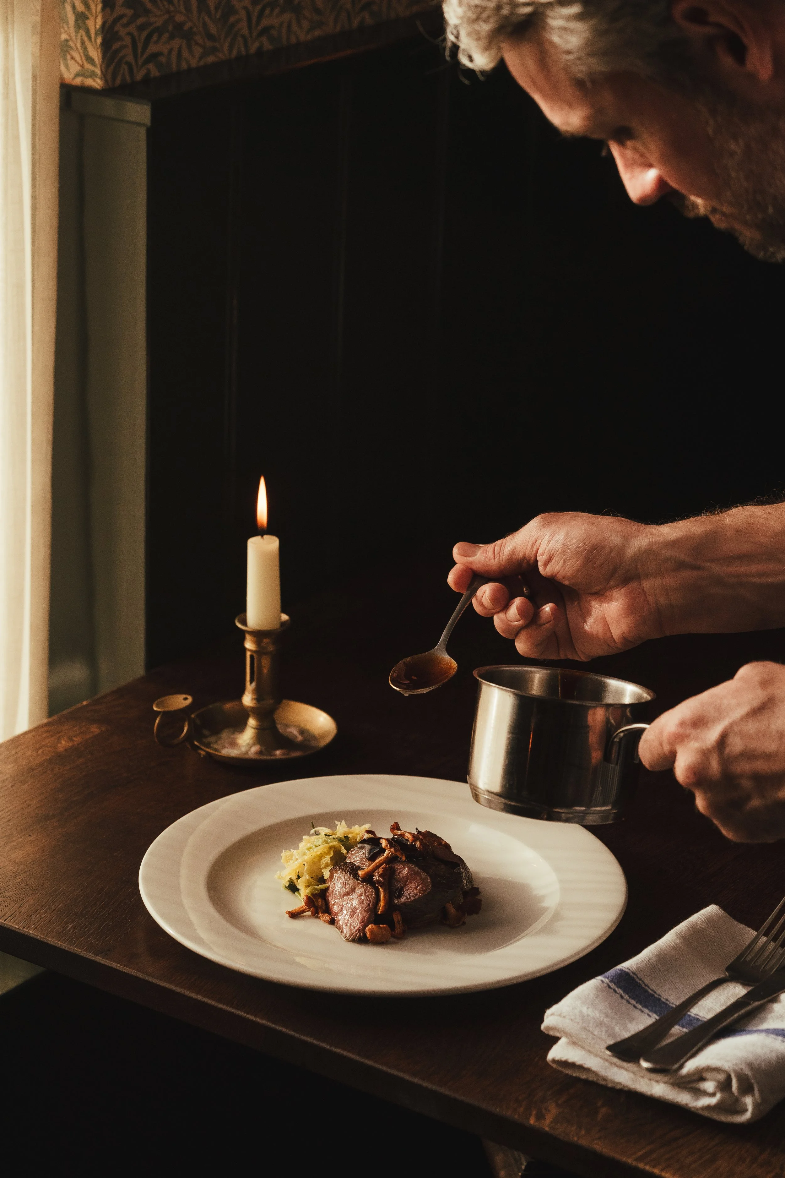 Man pouring gravy over cooked beef with mashed potatoes on a white plate, lit candle on table, at dinner setting.