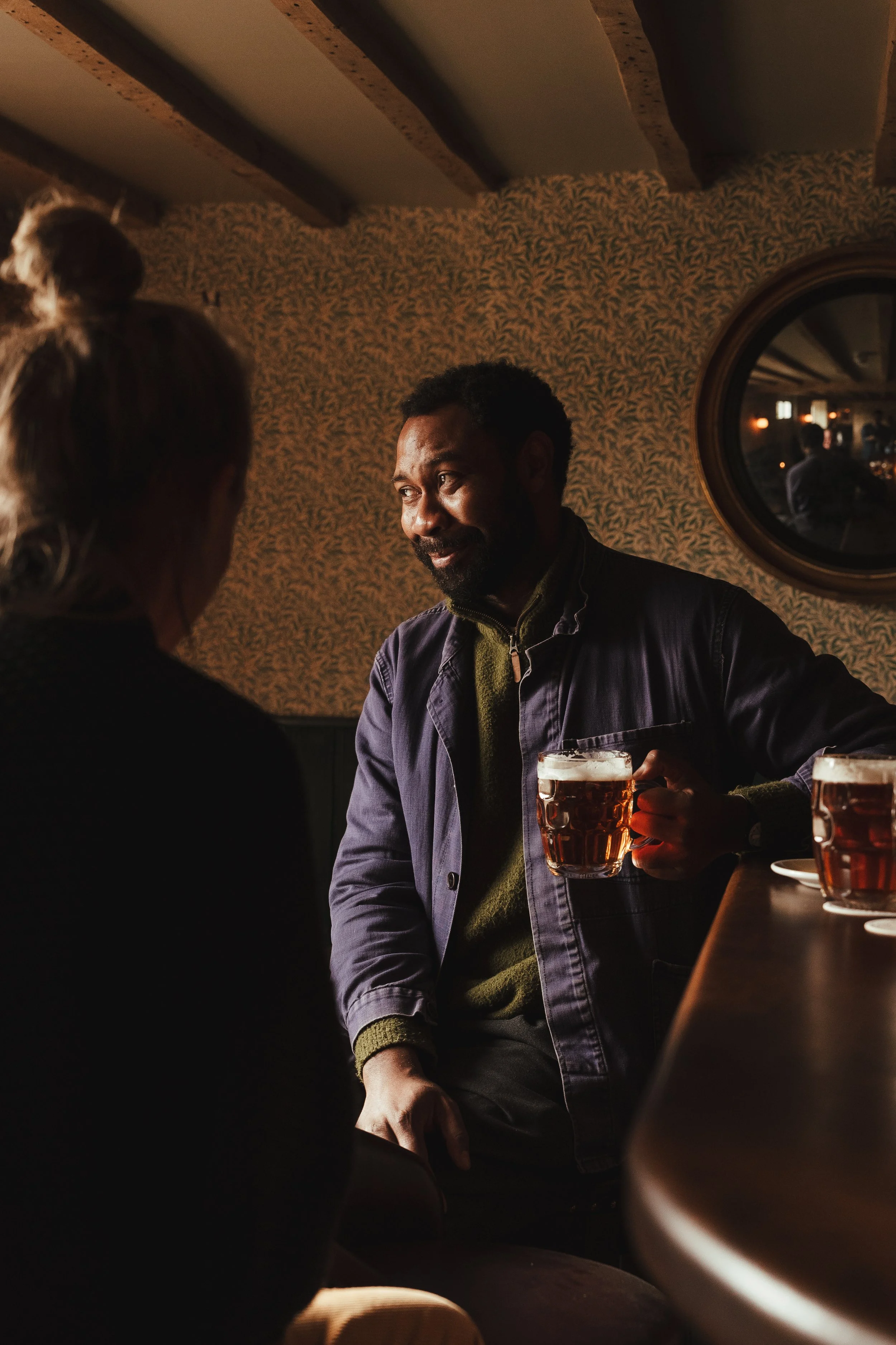 Two people having a conversation at a bar, one holding a beer mug, with dark wood and vintage wallpaper, in a dimly lit setting.