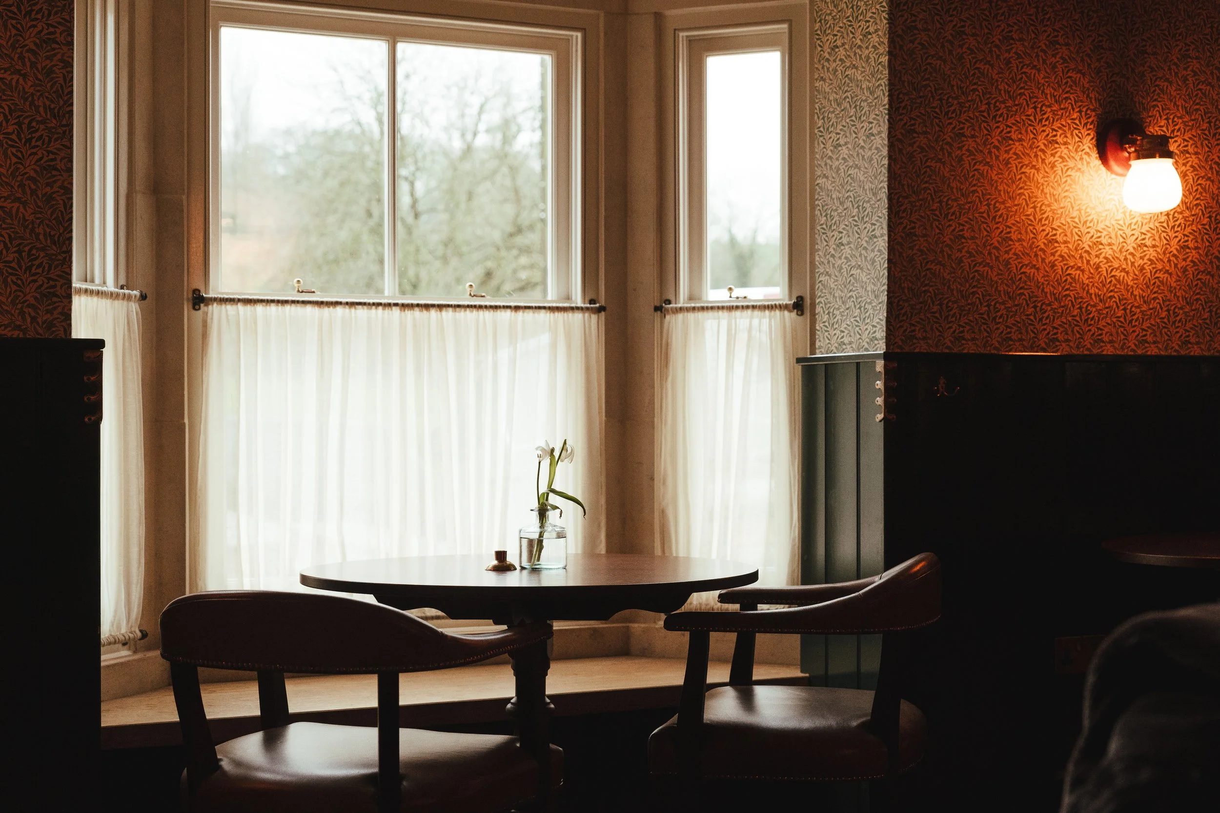 A cozy corner with a round wooden table and two leather chairs in front of large windows with cream curtains, decorated with a small glass vase containing a white flower, inside a softly lit room with floral-patterned wallpaper and a warm wall-mounte