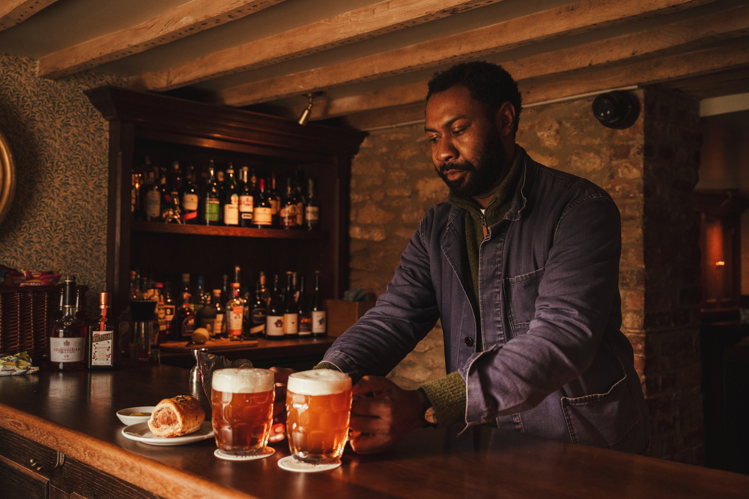A bartender pouring two pints of beer on a bar counter, with a plate of a pastry and a small dish nearby. The bar has shelves with liquor bottles and a rustic brick wall background.