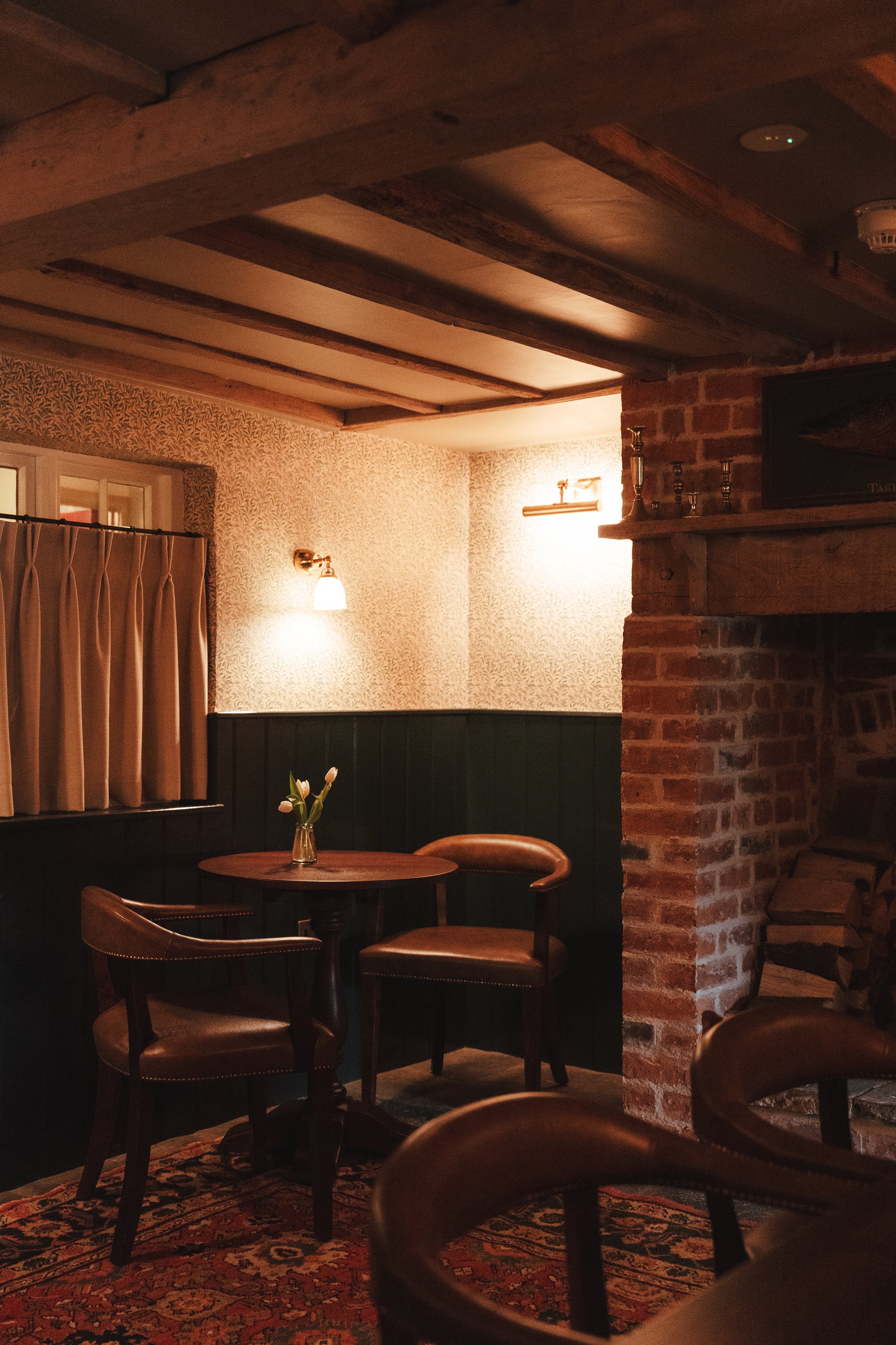A cozy dining area with a round wooden table, two leather chairs, and a vase of tulips, illuminated by warm lighting in a rustic interior.