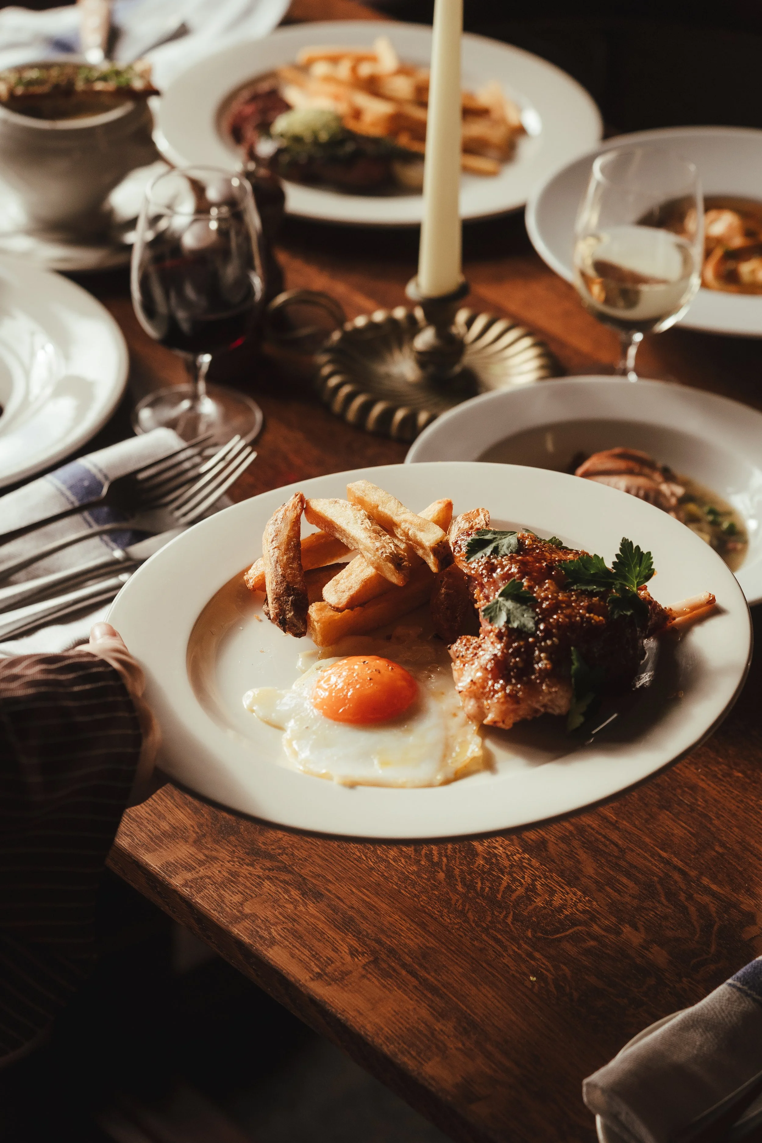 A dinner plate with fried egg, French fries, and a piece of cooked meat, likely lamb, garnished with herbs. The table is set with silverware, wine glasses, bowls of soup, and other plates of food.