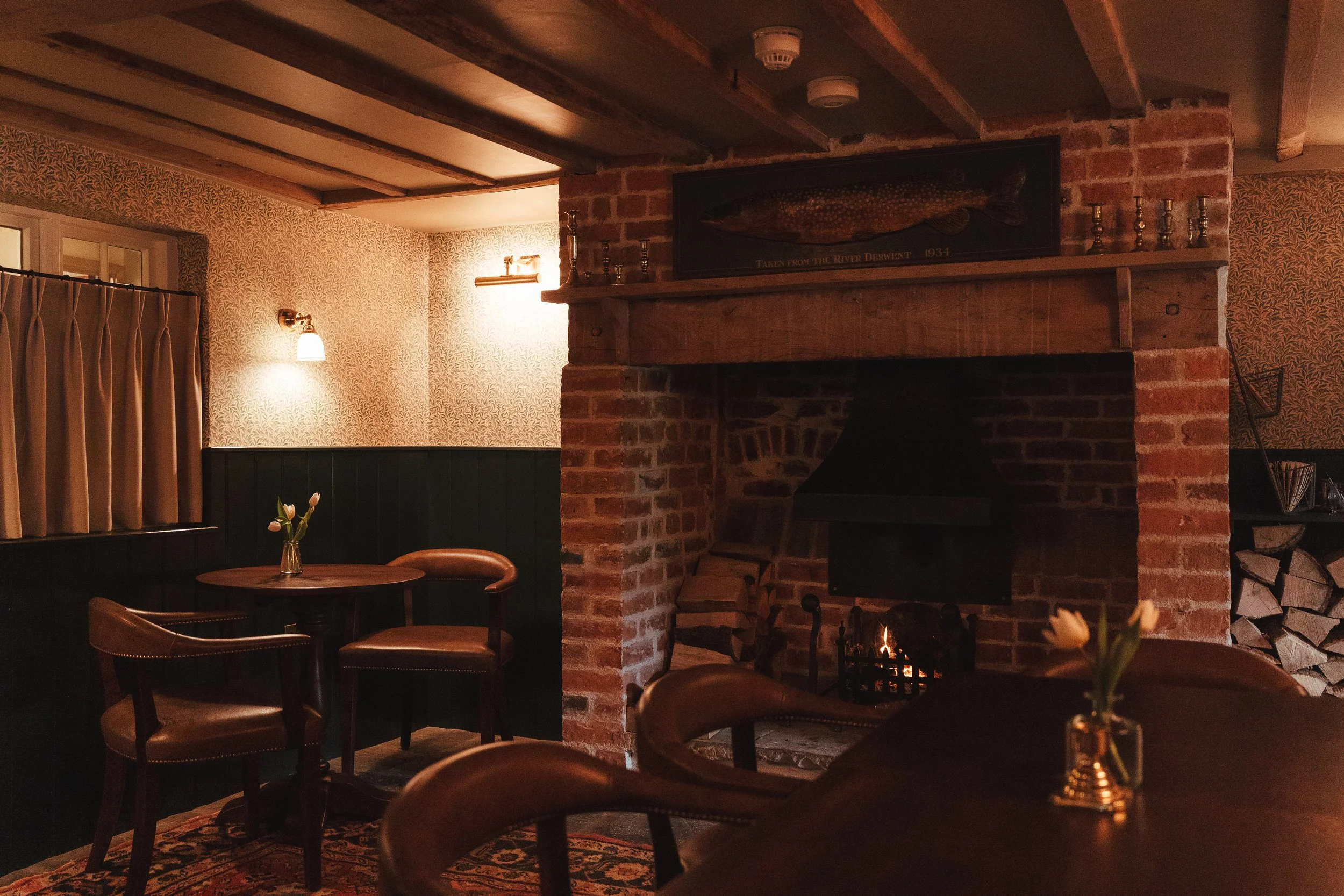 Cozy dining room with a brick fireplace, wood logs, small tables, leather chairs, and wall-mounted lights, decorated with a framed fish display and flowers on the tables.