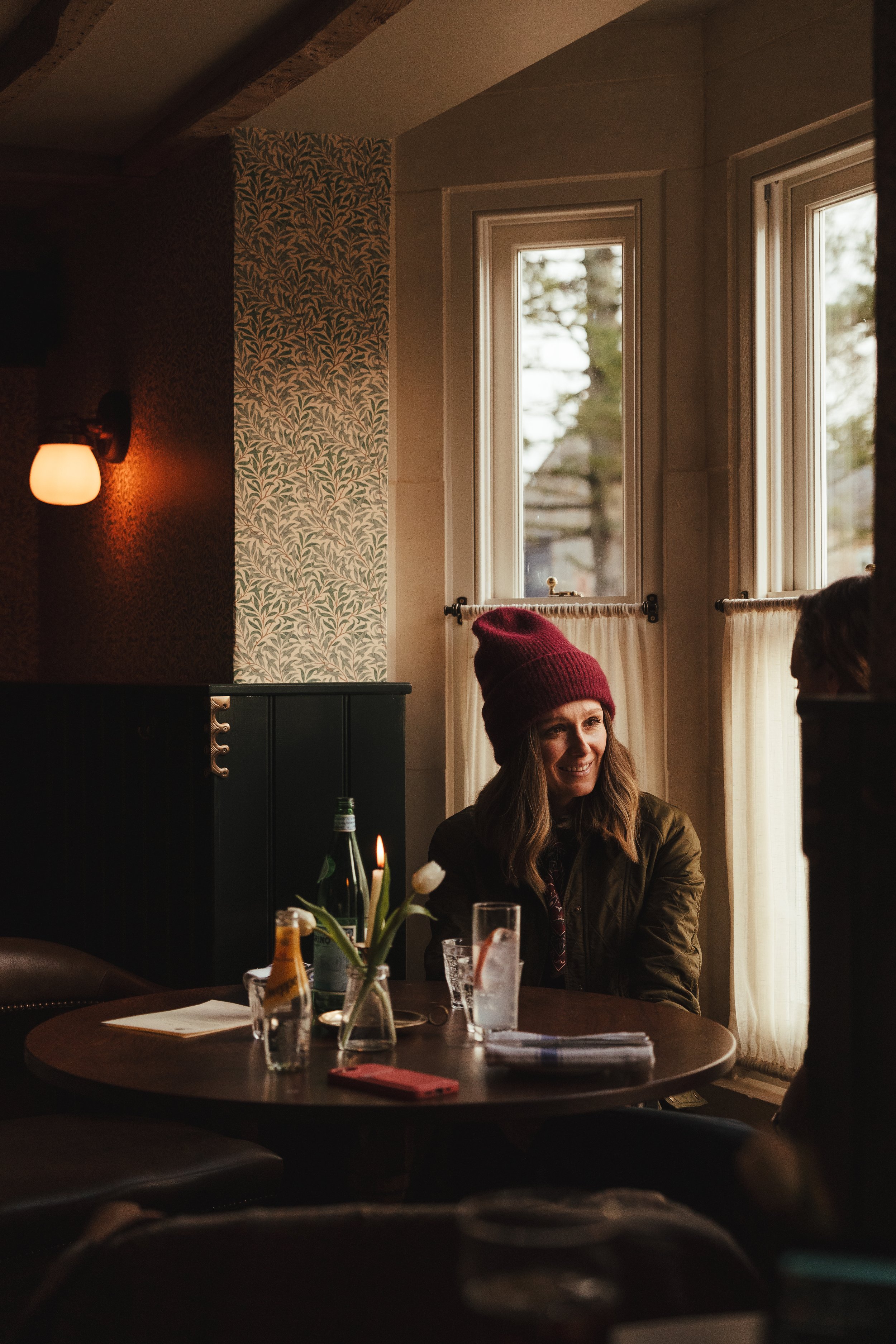 A woman wearing a red beanie sitting at a table in a cozy cafe or restaurant, with a window behind her and a vase with white flowers on the table.