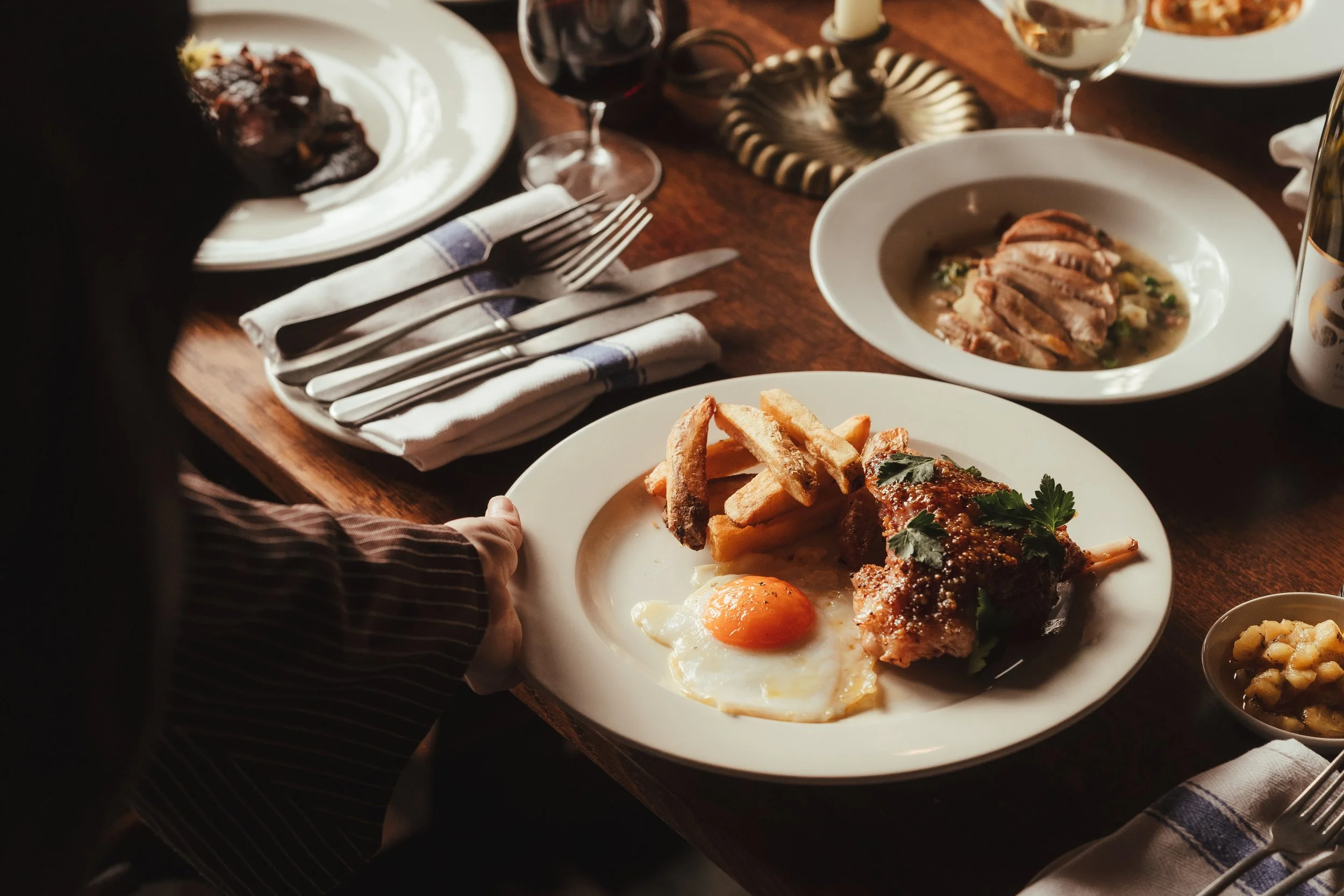 A plate with French fries, a sunny side up egg, and a piece of cooked meat garnished with herbs. A person's hand is holding the plate. On the table, there are additional plates with food, wine glasses, cutlery, and napkins.
