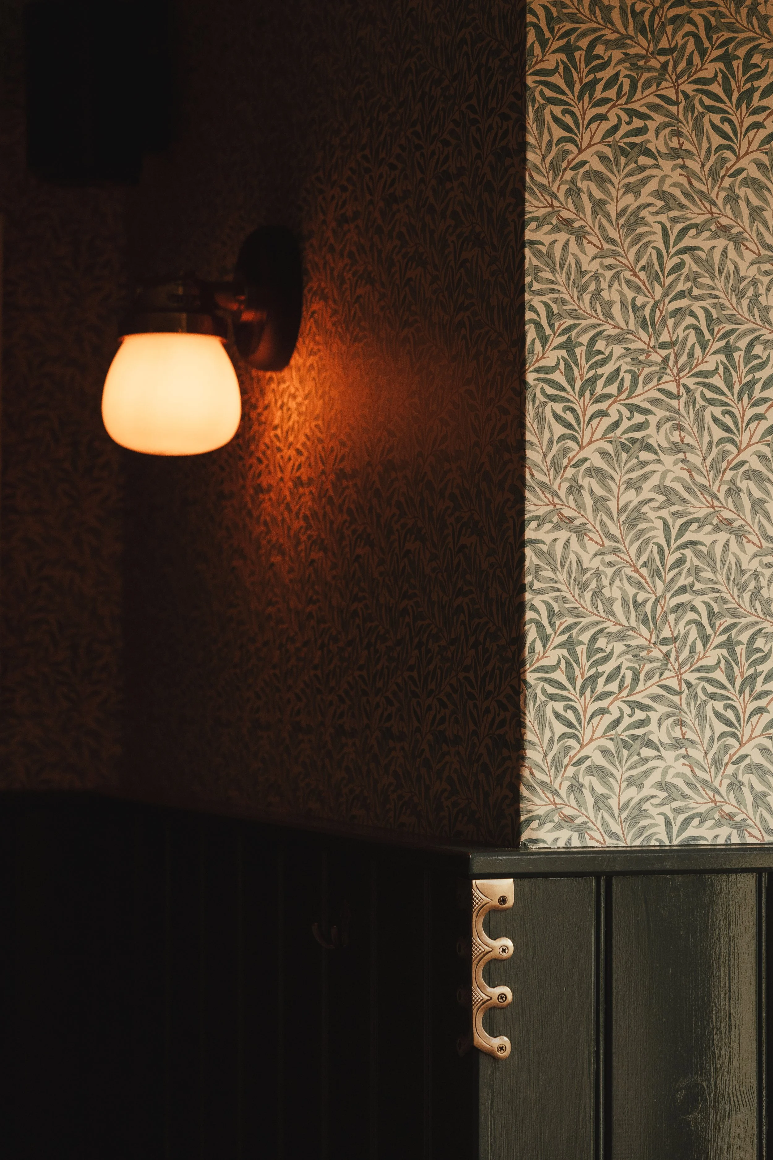 Interior corner with a wall-mounted light fixture emitting warm light, dark patterned wallpaper on one wall, and light-colored wallpaper with leafy pattern on the adjacent wall, along with dark wood paneling.