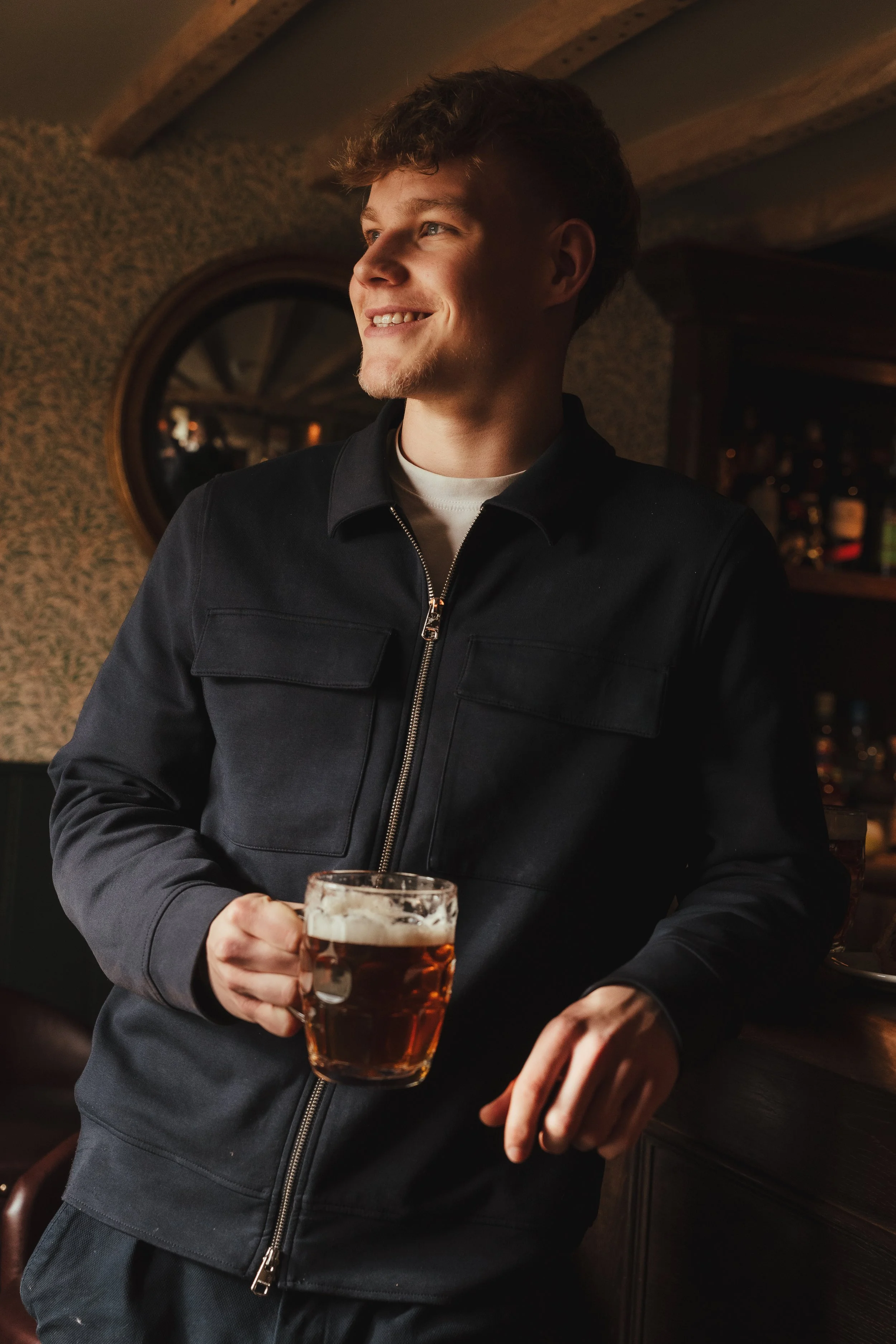 A young man with curly hair, smiling, holding a glass of beer in a bar or pub setting.