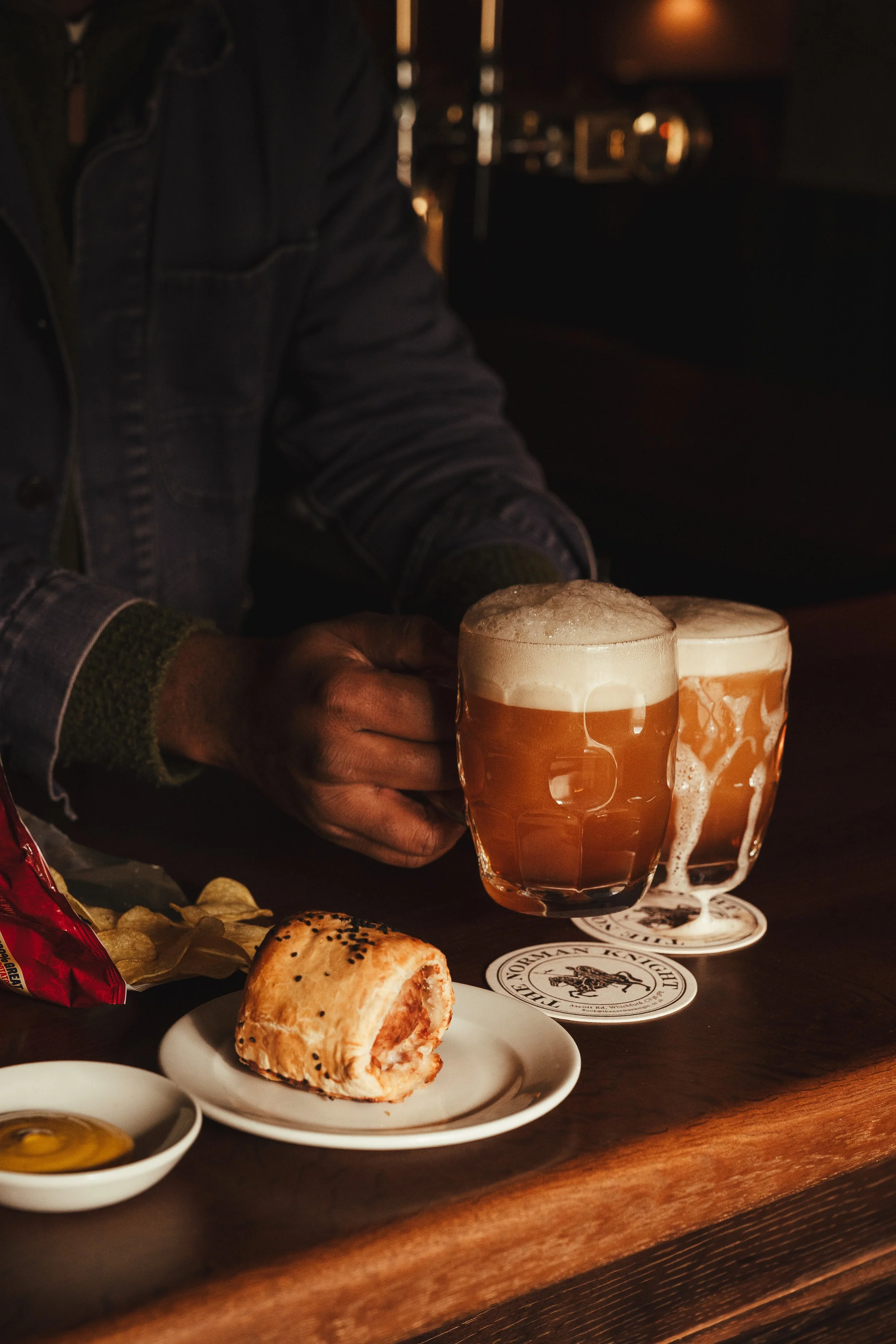 A person holding two glasses of beer on a wooden bar counter, with a plate of a half-eaten pastry and a small dish of honey or syrup nearby.