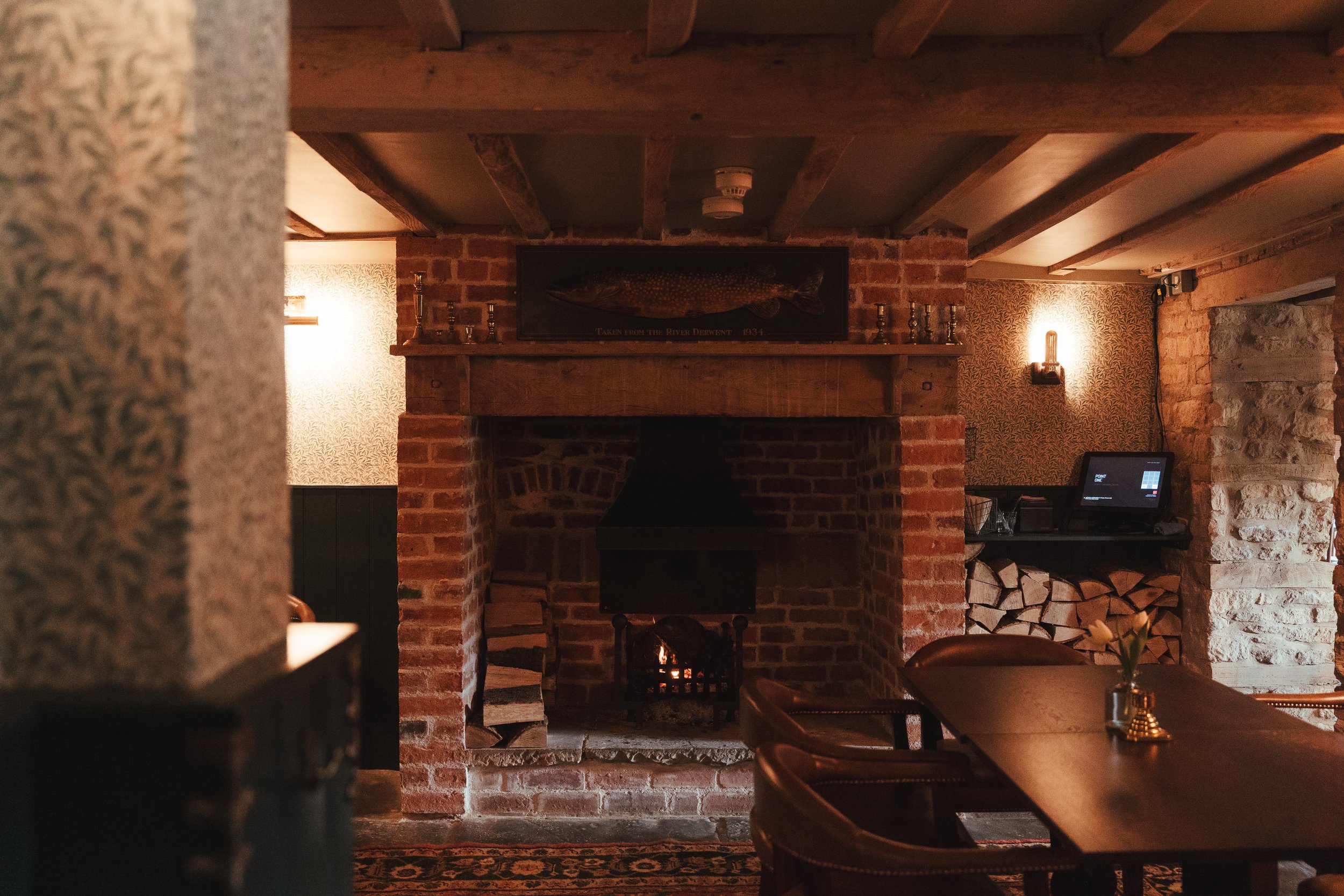 Cozy restaurant interior with a brick fireplace, lit with a small fire, with a framed fish painting above it. The room has wooden beams on the ceiling and warm lighting, with a table and chairs in the foreground and a wood storage area to the right.
