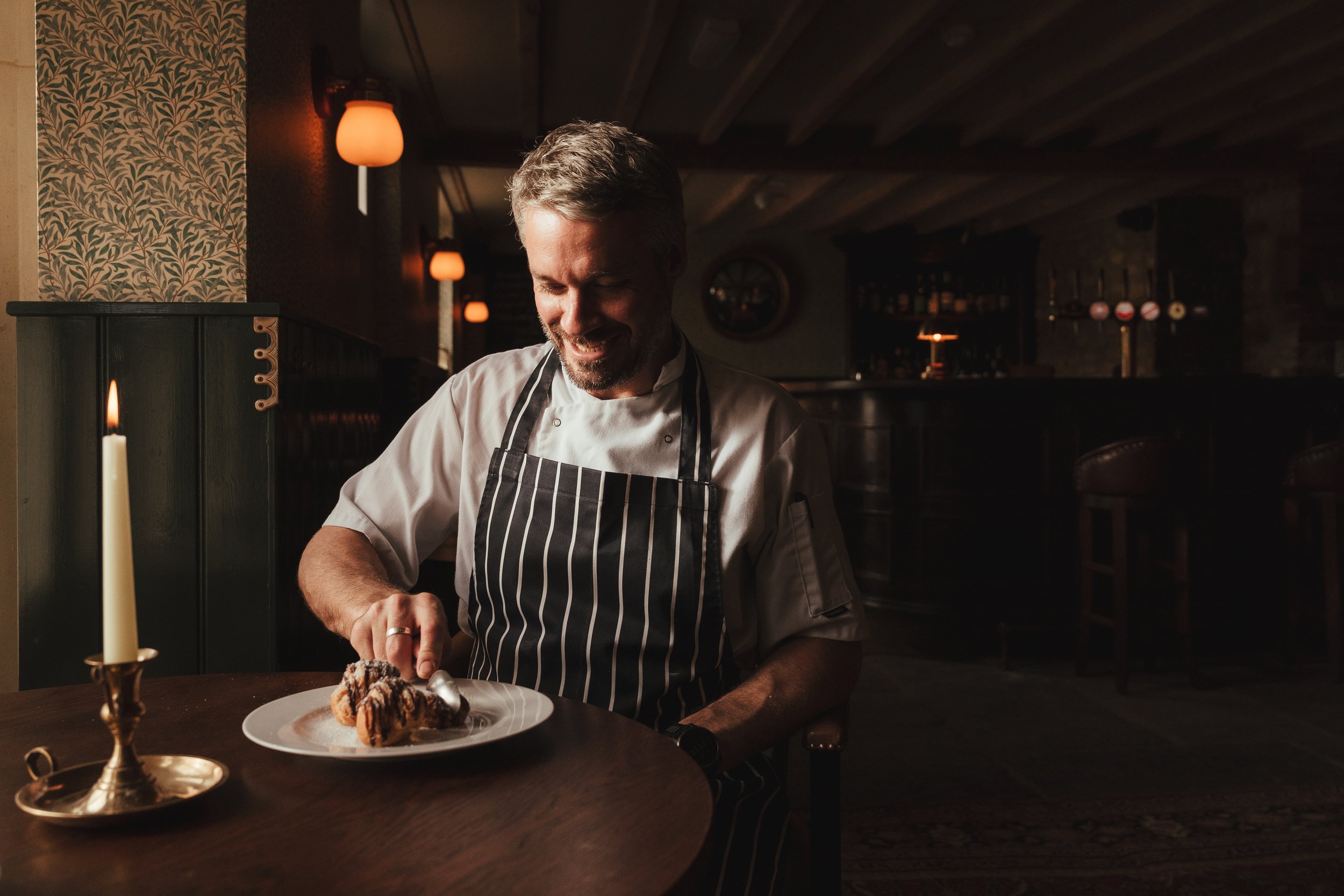 A chef in a striped apron is smiling while preparing a dessert on a white plate in a dimly lit restaurant, with a single lit candle on the table.