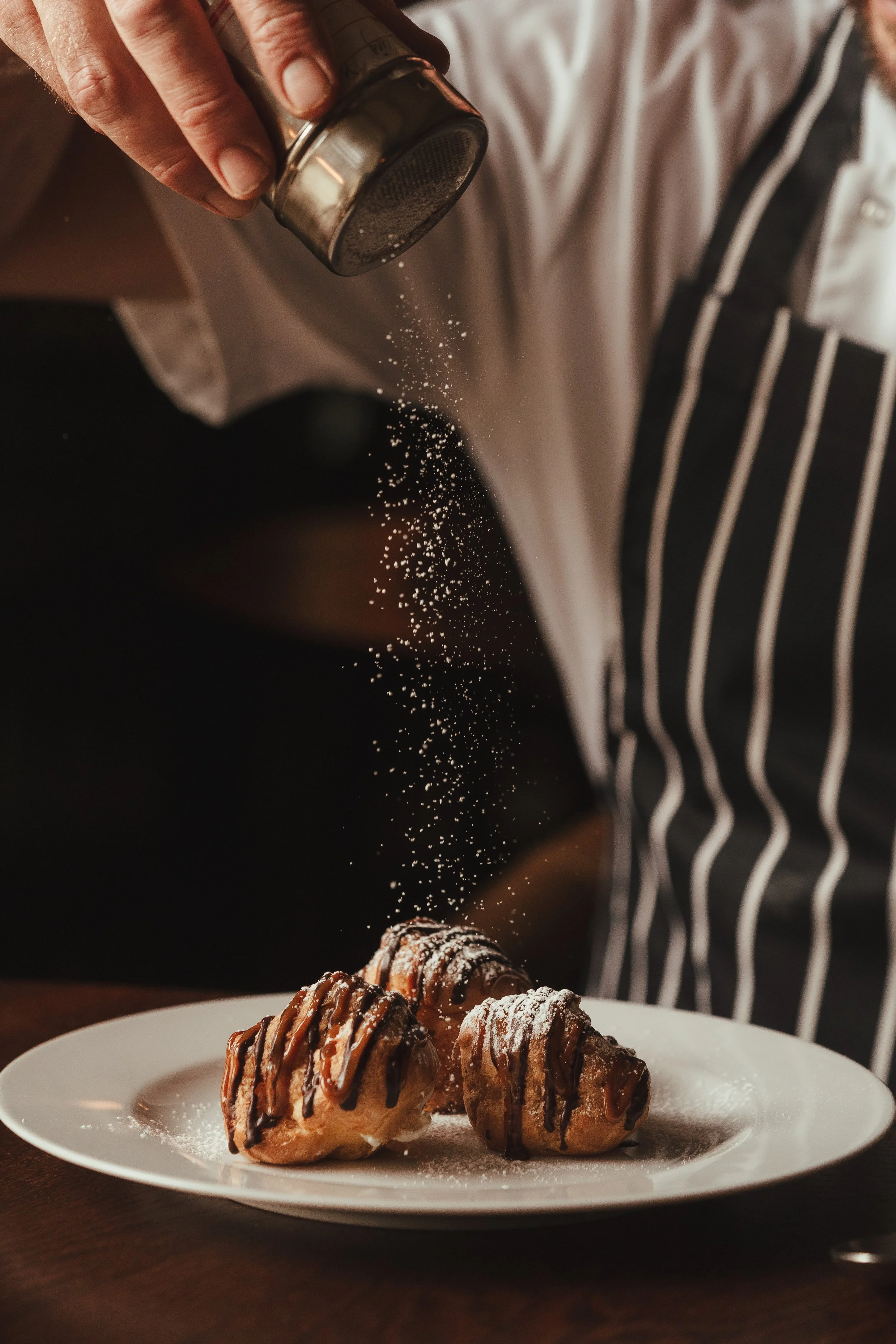A person wearing a black and white striped apron sprinkling powdered sugar onto three profiteroles drizzled with chocolate sauce on a white plate.