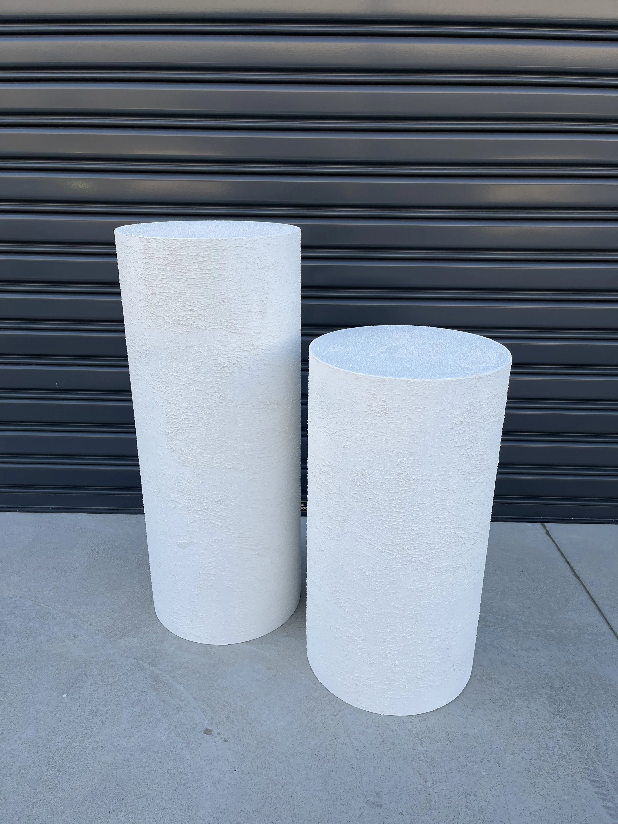 Two white cylindrical pedestals with textured surfaces, placed outdoors on a concrete surface in front of a black metal rolling door.
