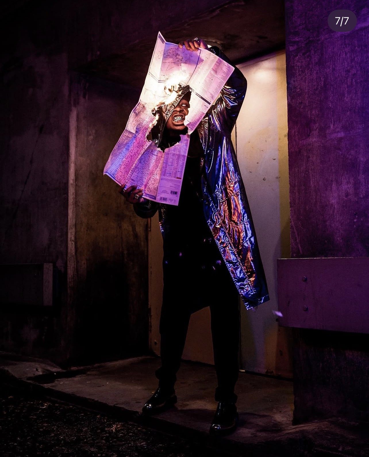 A person wearing a black outfit and shiny jacket holding a torn piece of paper or newspaper in an industrial-style setting with purple and yellow lighting.