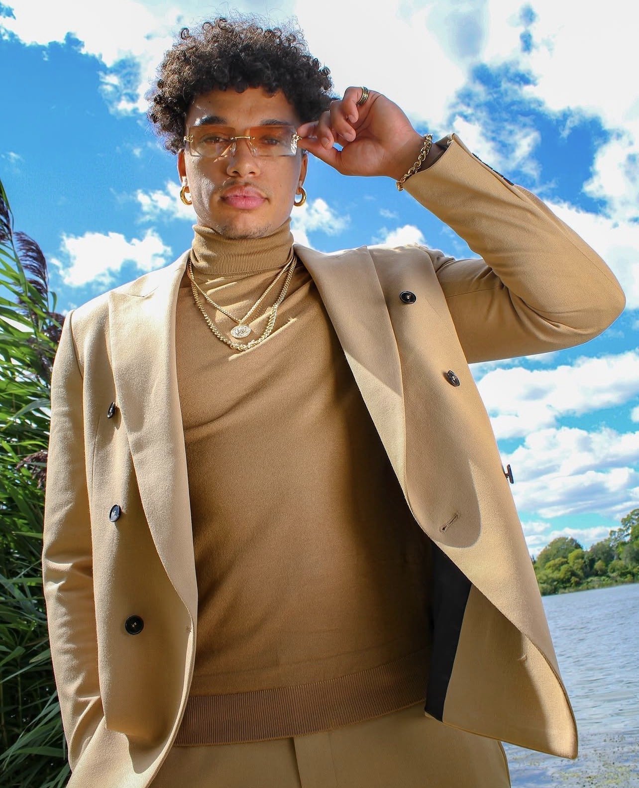 Man wearing glasses, gold jewelry, and a tan suit with a river and cloudy sky in the background.