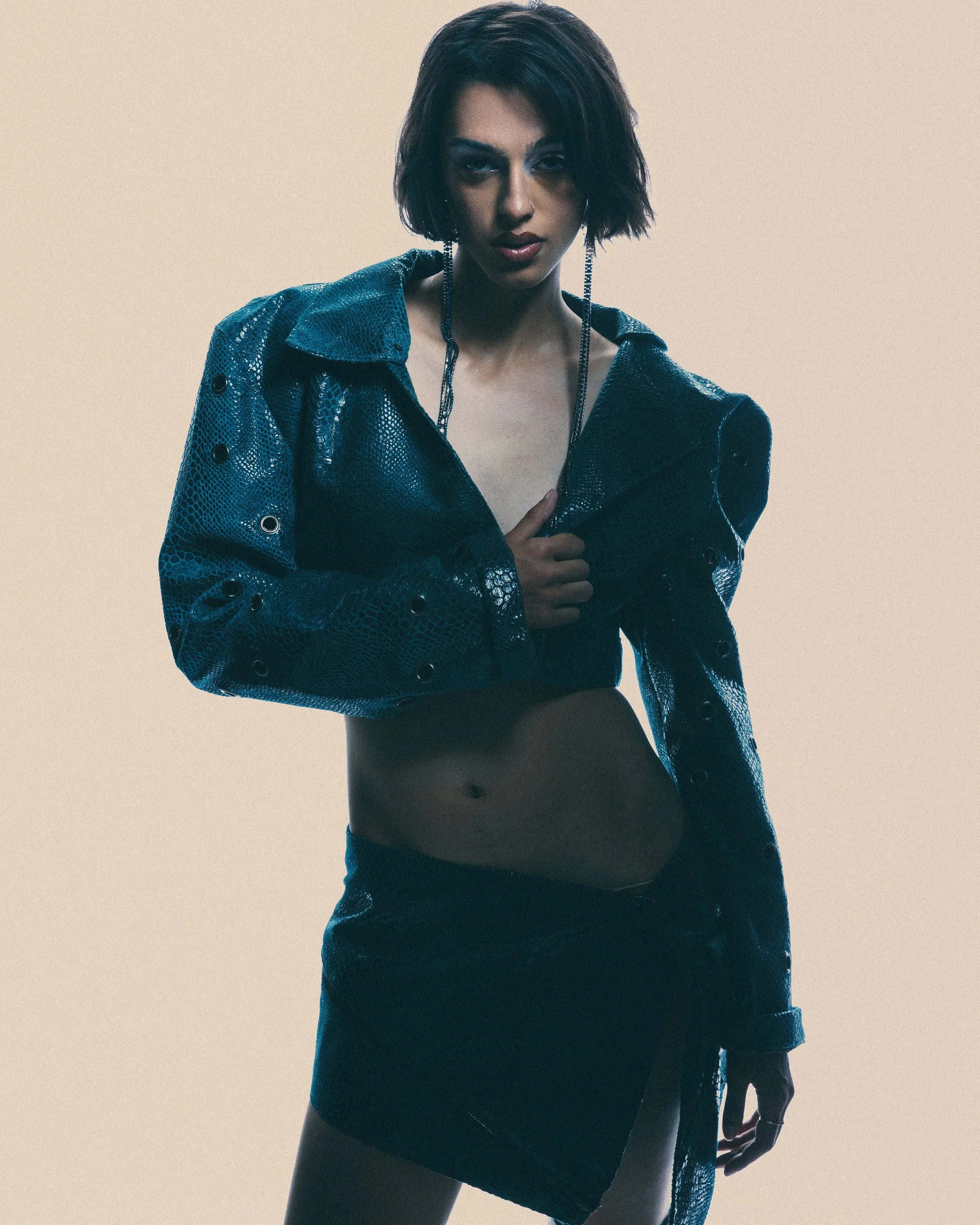 A young woman with dark hair and makeup, wearing a shiny blue jacket and a black skirt, standing against a plain background.