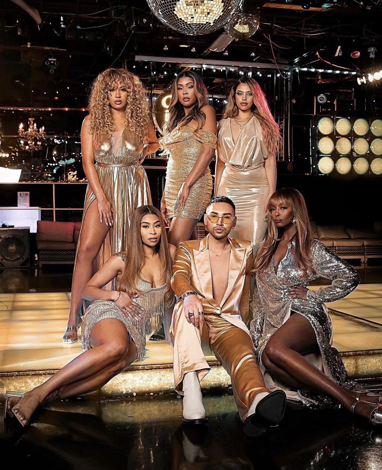Group of six fashionable people dressed in gold and metallic clothes posing on a dance floor with a disco ball overhead in a nightclub.