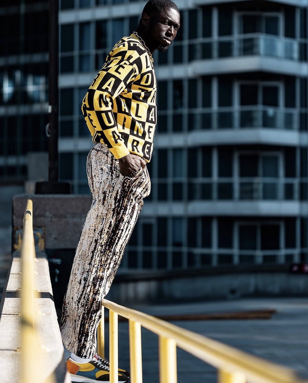 A man standing outdoors on a ledge with yellow railings in front of a modern glass building, wearing a yellow and black patterned sweater, baggy printed pants, and colorful sneakers.