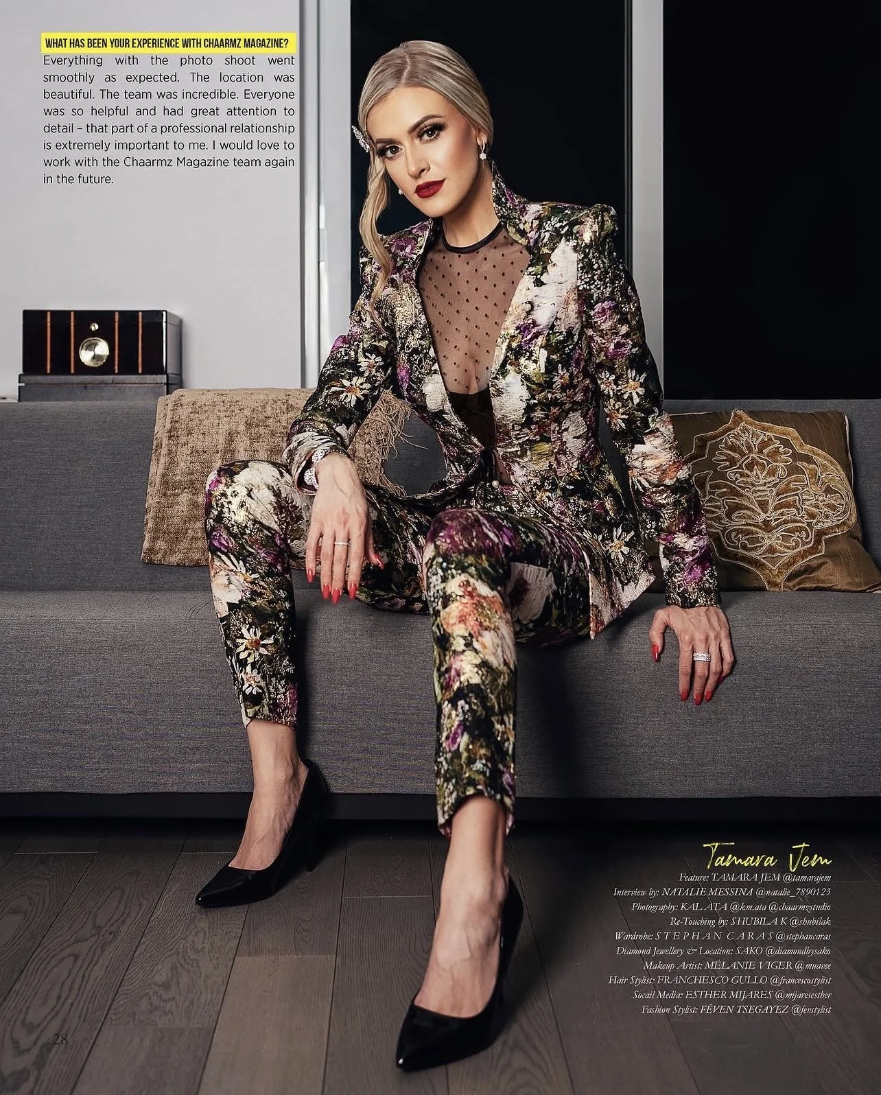 A woman in a floral suit with a sheer black polka dot top sitting on a gray couch with decorative pillows, wearing black high heels, and posing for a photoshoot with a professional setting.