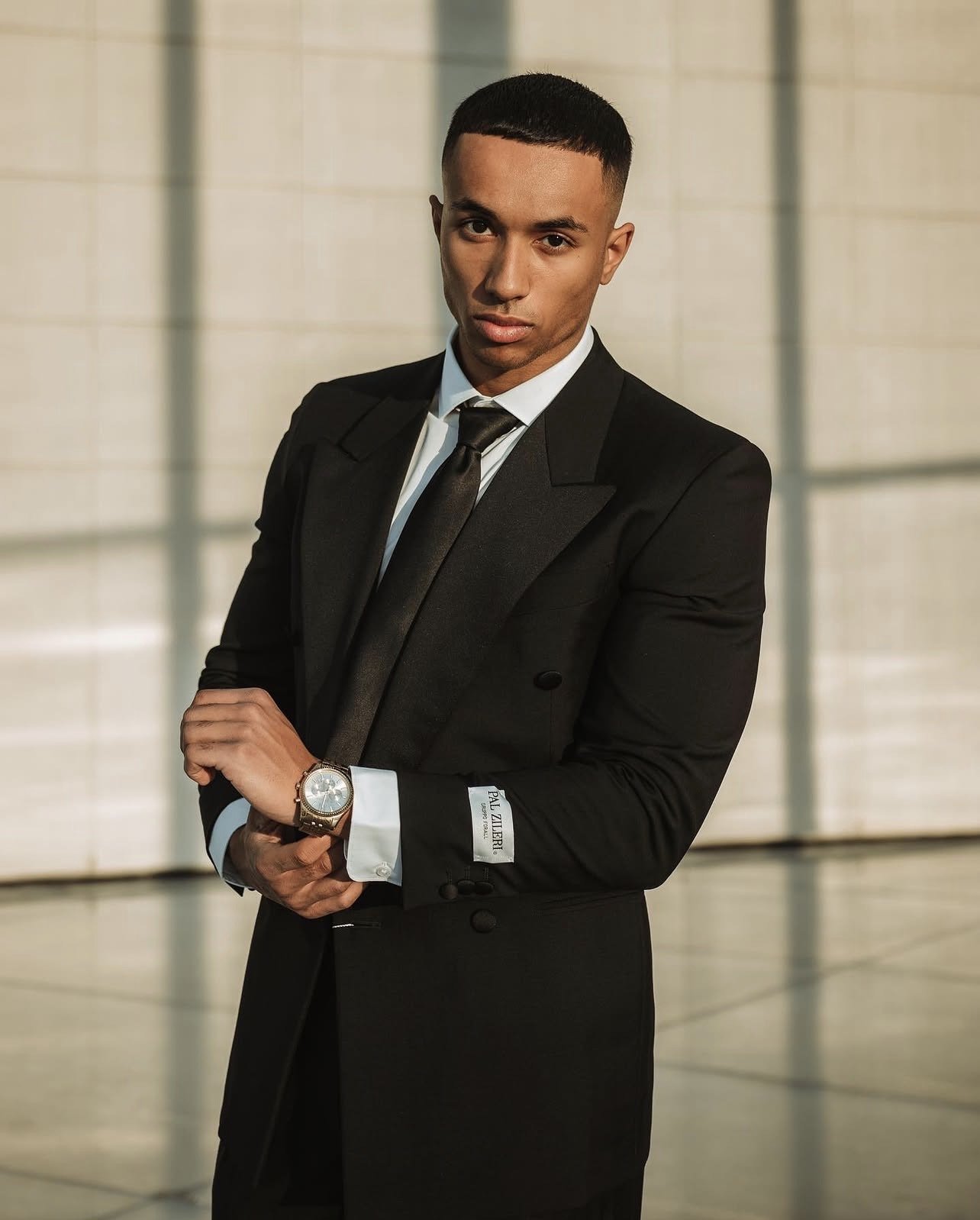 A young man in a black tuxedo with a satin lapel, white dress shirt, and black tie, standing indoors near a large window with sunlight casting shadows.