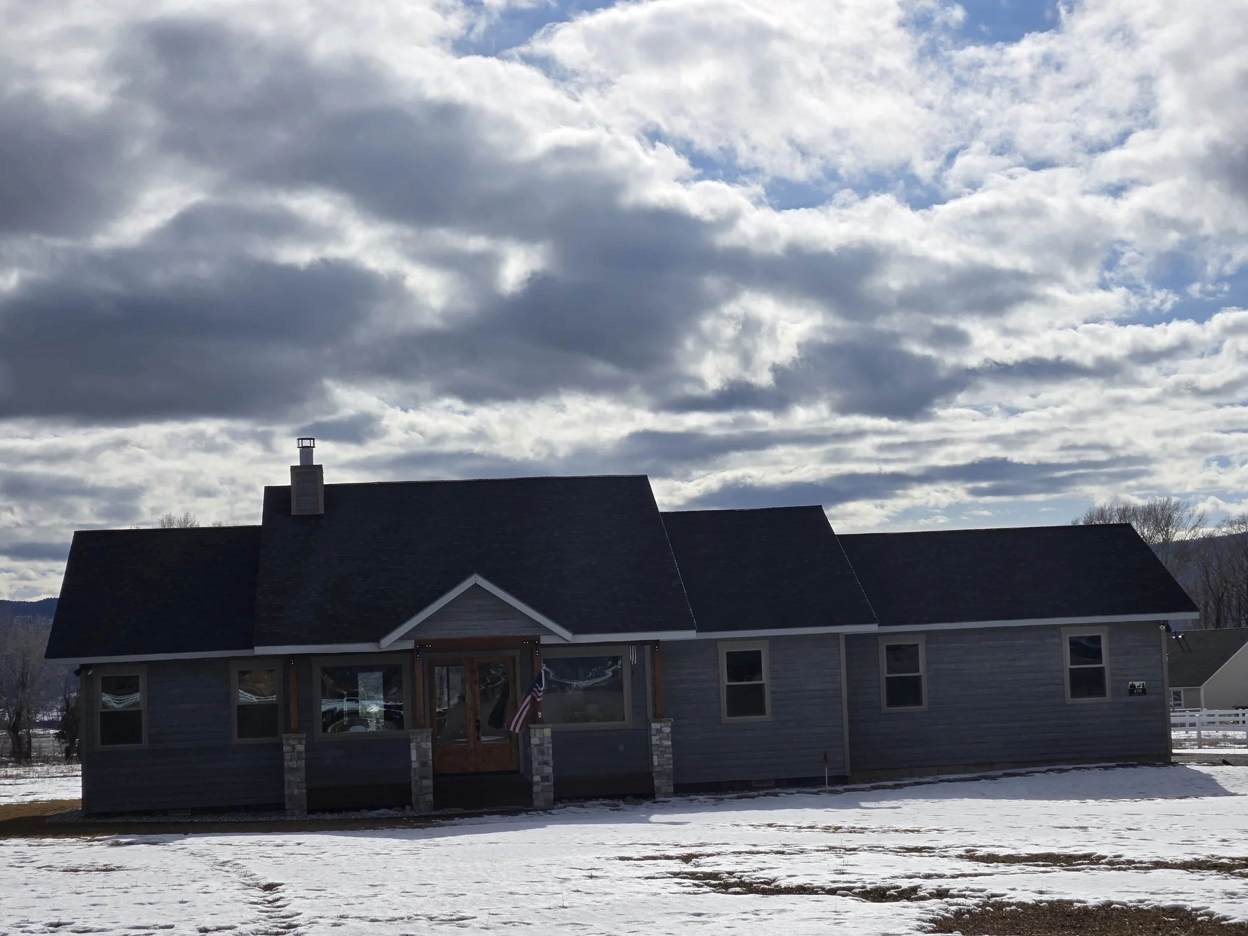 A single-story house with grey siding, dark roof, and a front porch with stone pillars, set against a partly cloudy sky and patches of snow on the ground.
