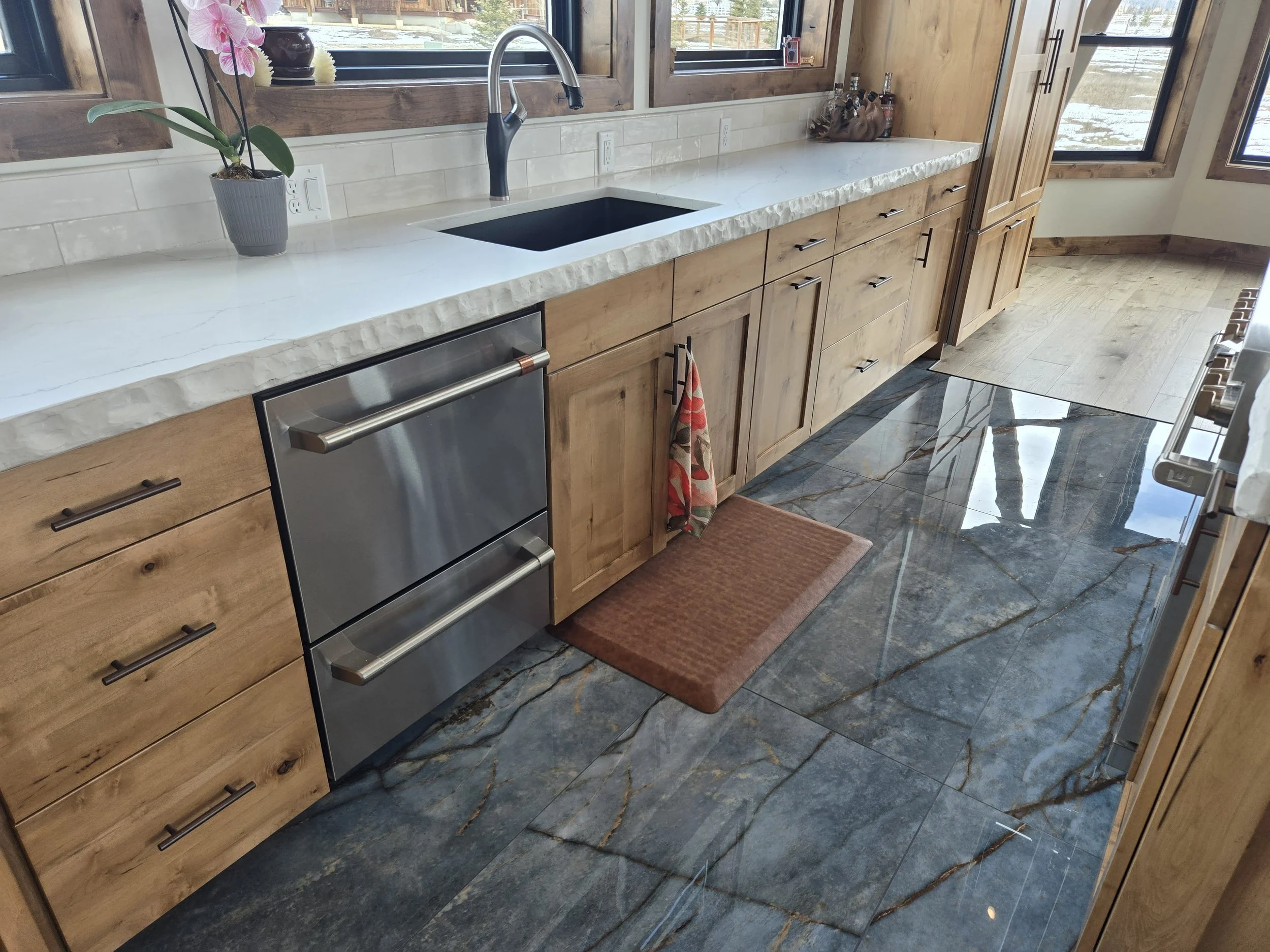 Kitchen with wooden cabinets, white marble countertop, black farmhouse sink, stainless steel double oven, and large windows. There is a potted orchid plant on the counter and colorful dish towels hanging. The floor is polished dark marble with gold v