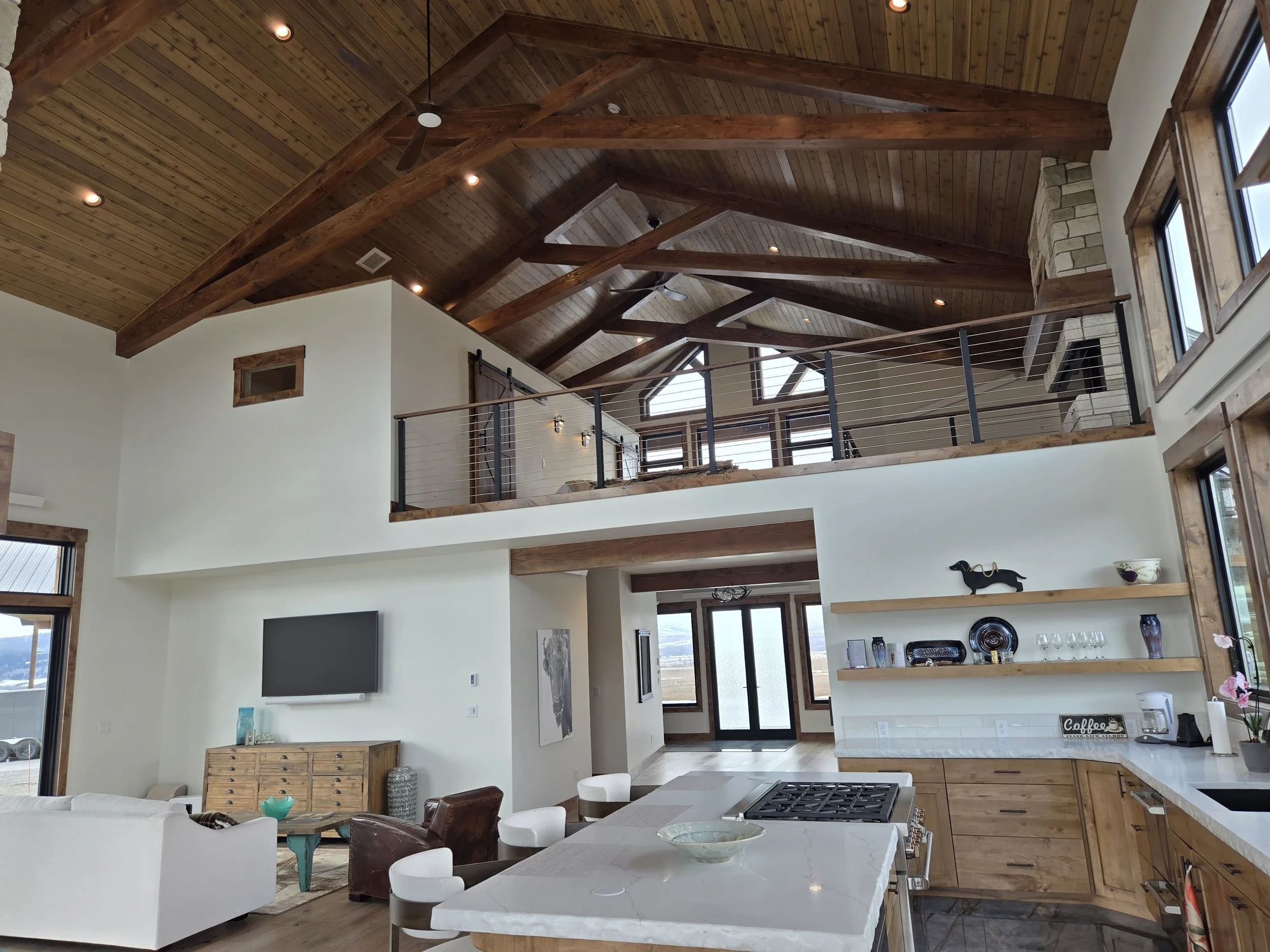 Interior of a modern house with a high vaulted wooden ceiling, exposed beams, large windows, an open kitchen with a white island, and a living area with a wall-mounted TV.
