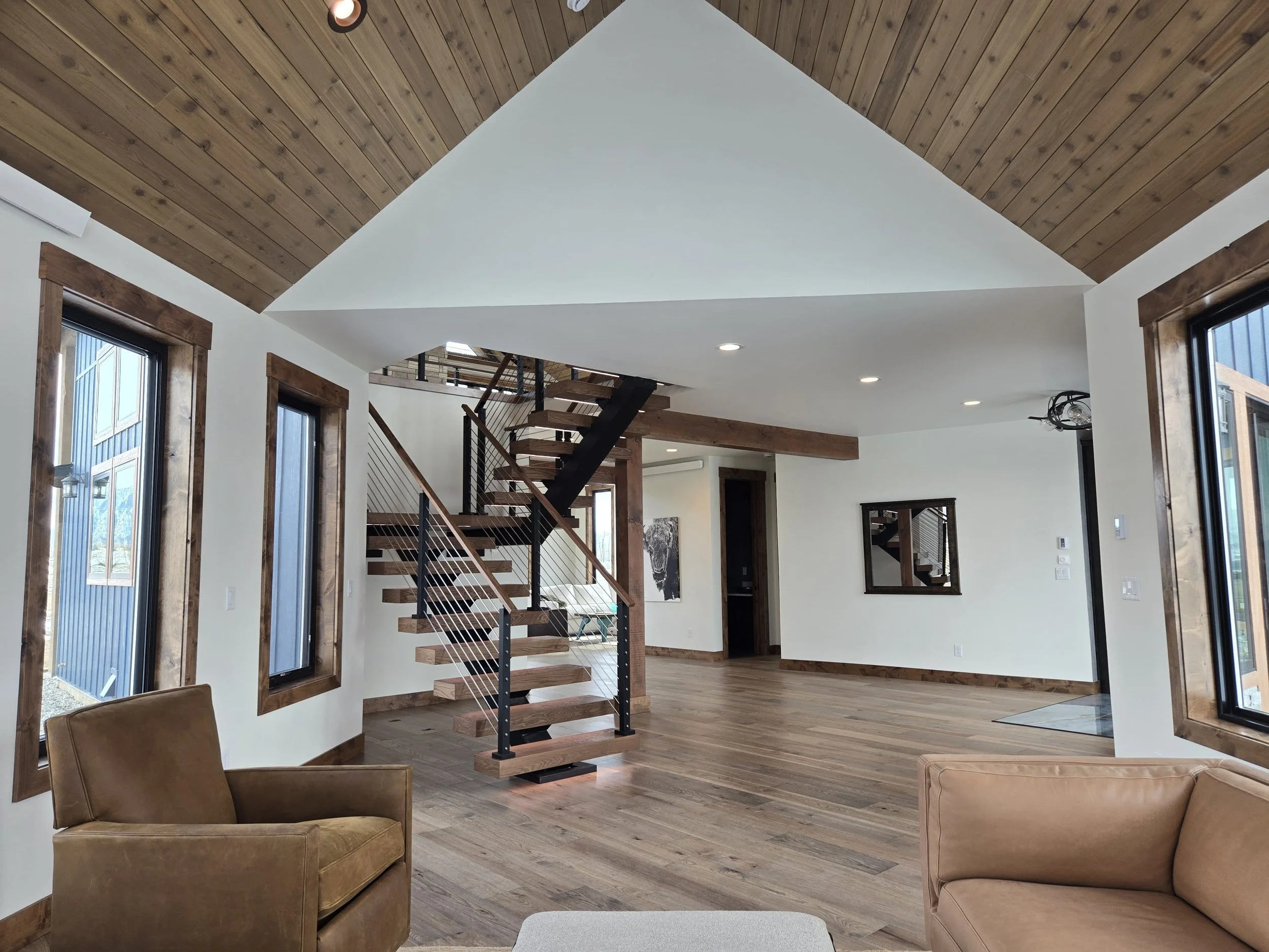 Interior of a modern home living room with wood floors, white walls, large windows with wood trim, a wooden ceiling, a staircase with wooden steps and metal railing, and beige armchairs.