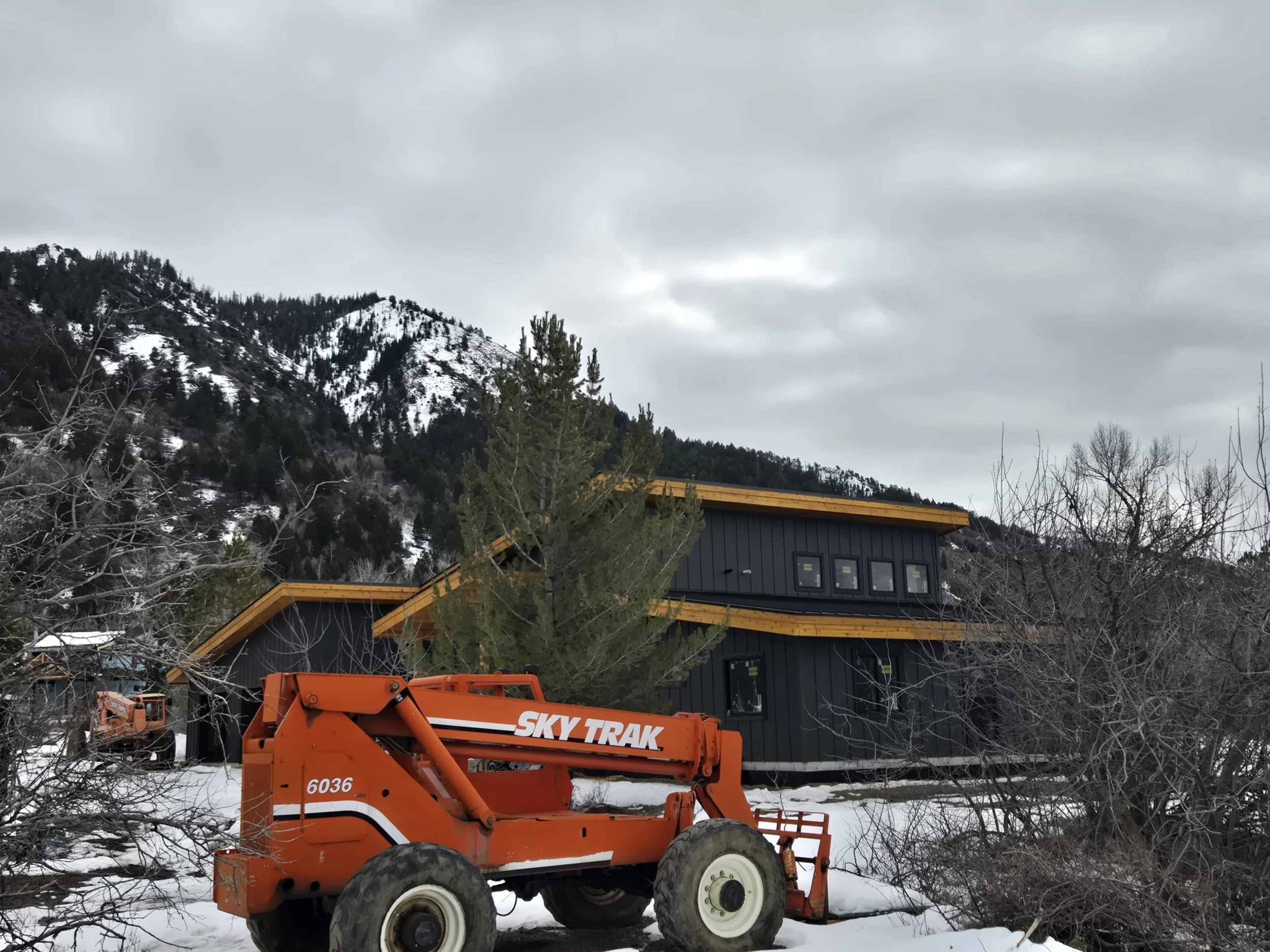 A modern black house with yellow accents in a snowy landscape, surrounded by leafless trees and mountains in the background, with an orange SkyTrak construction vehicle nearby.