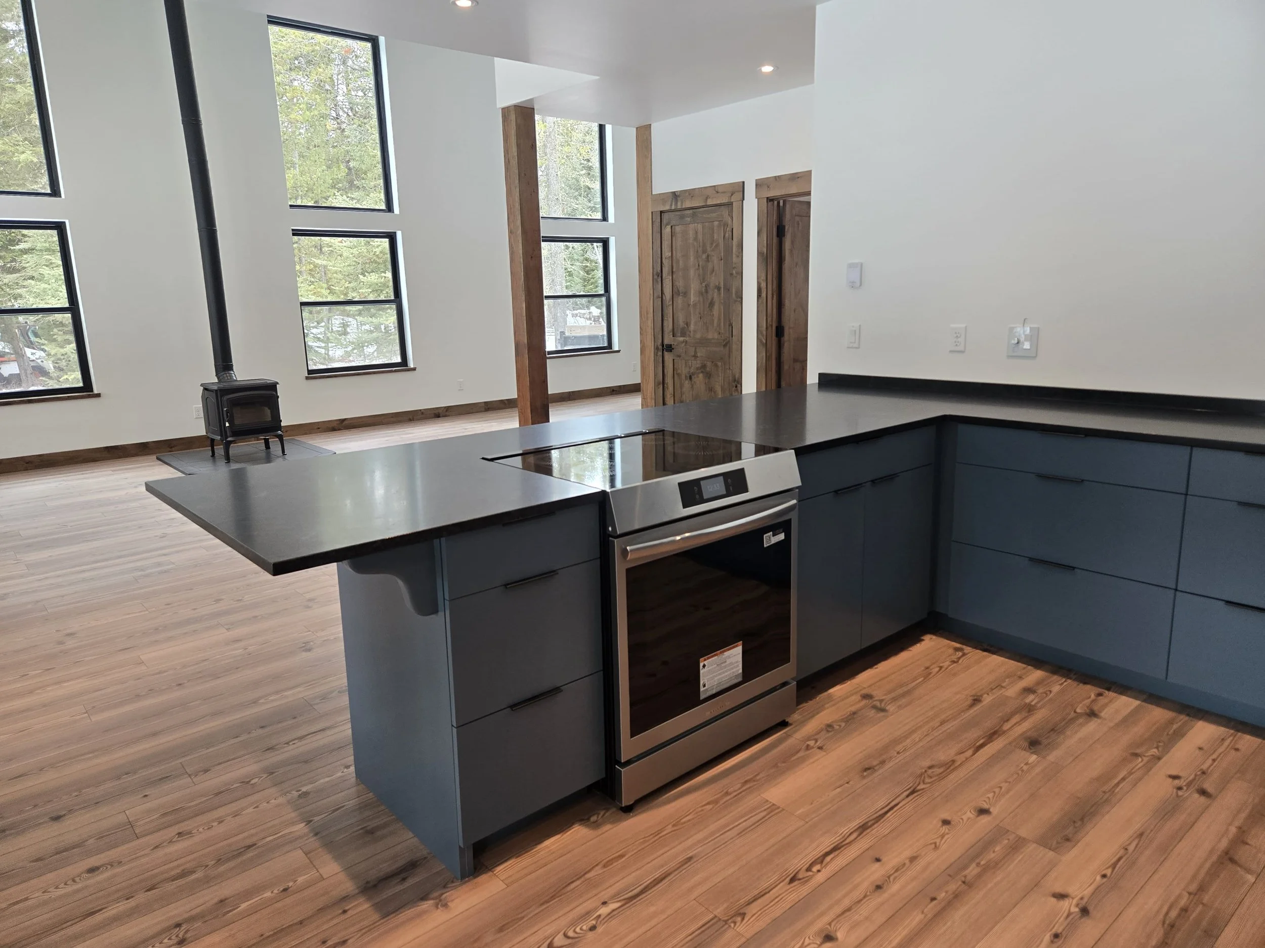 Modern open-concept kitchen with black countertops, stainless steel oven, blue cabinetry, wooden floors, and large windows in the living space with a black wood stove.