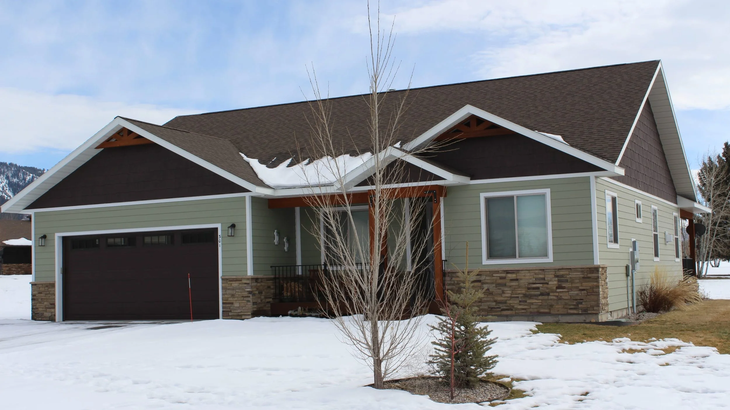 Modern house with a brown roof, green siding, stone accents, and a garage, set in a snowy landscape with leafless trees.