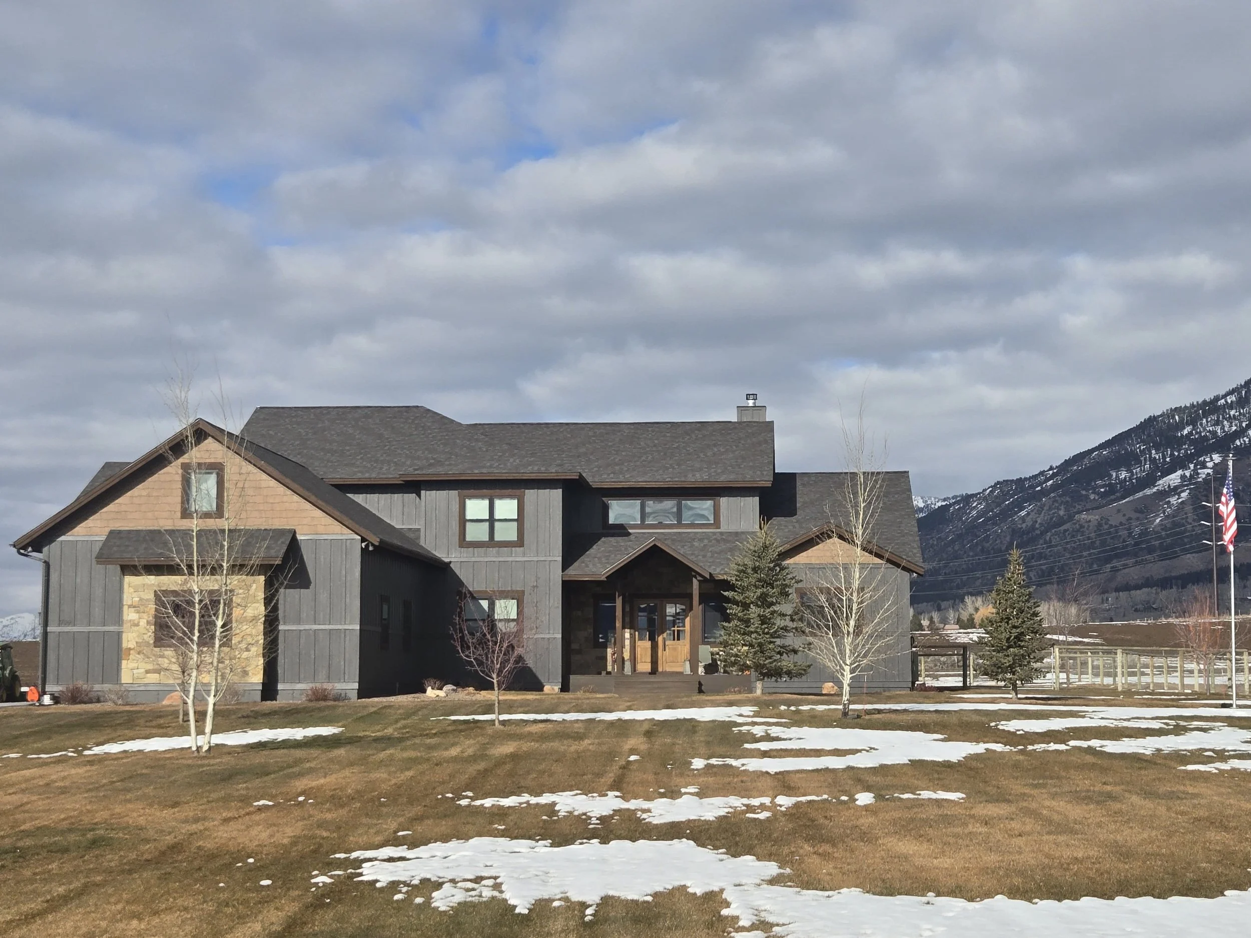 A large modern house with dark gray vertical siding and light brown accents, situated in a rural area with snow patches on the lawn, leafless trees, and mountains in the background under a partly cloudy sky.