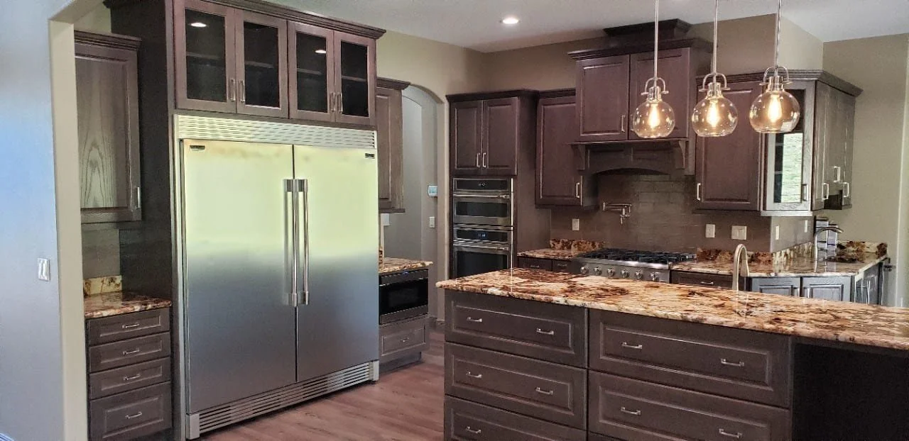 A modern kitchen with dark wood cabinets, granite countertops, stainless steel appliances including a double-door refrigerator, oven, microwave, and stovetop, and pendant lights hanging above the island.
