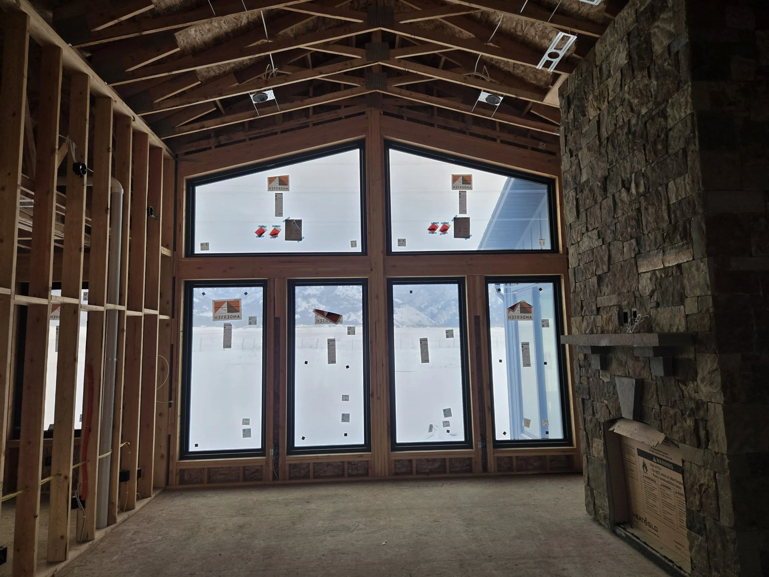 Interior view of a house under construction with large window panels, exposed wooden framing, and a stone fireplace. Outside the window, a snowy mountain landscape is visible.