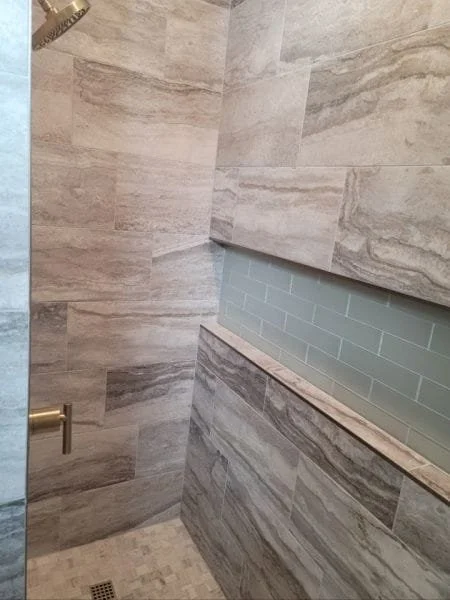 A shower wall with large beige and gray stone tiles and a partial glass partition with green subway tiles behind it.
