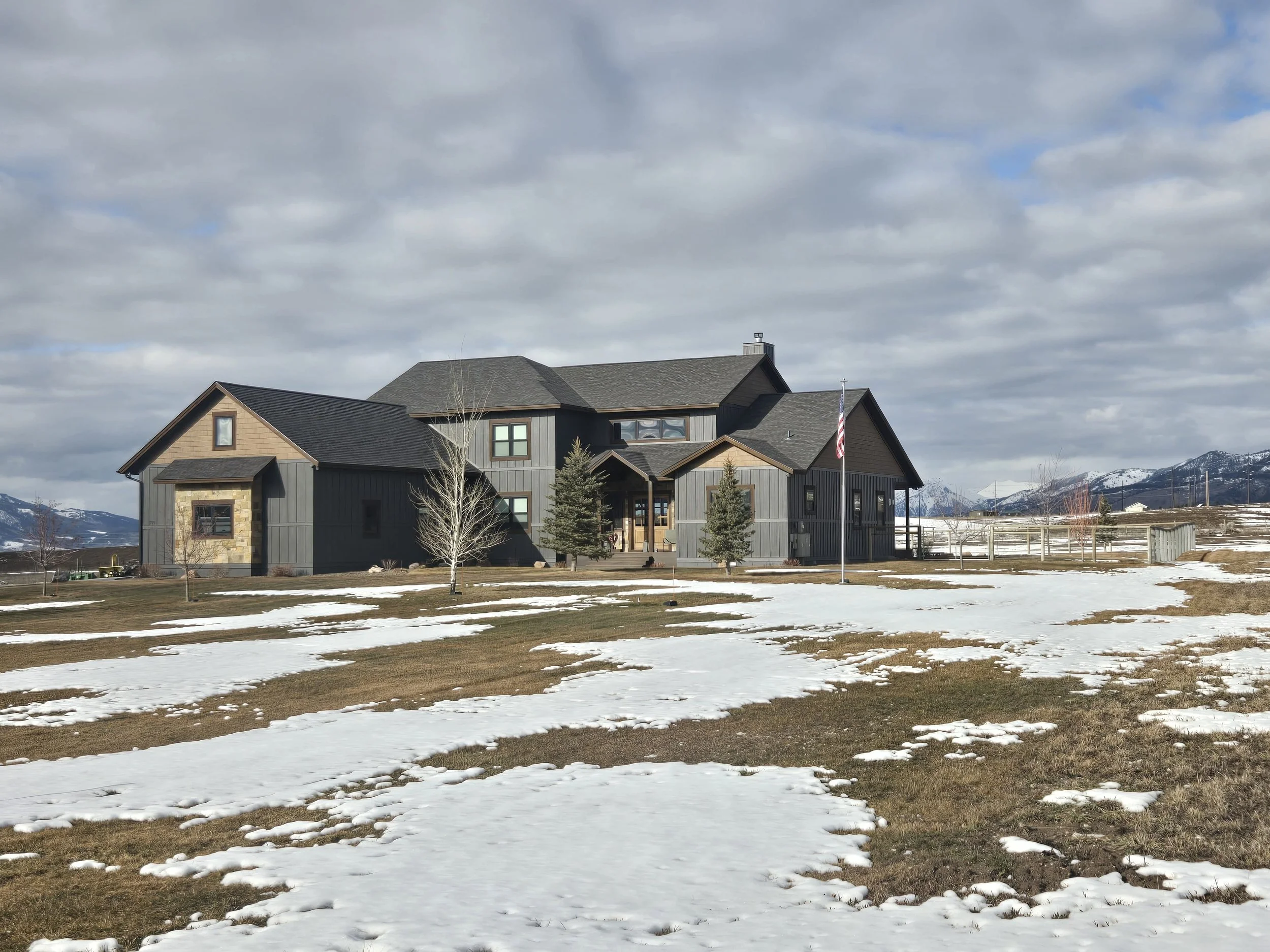 Large modern house with dark gray siding, surrounded by snow patches on the lawn, with mountains and cloudy sky in the background.