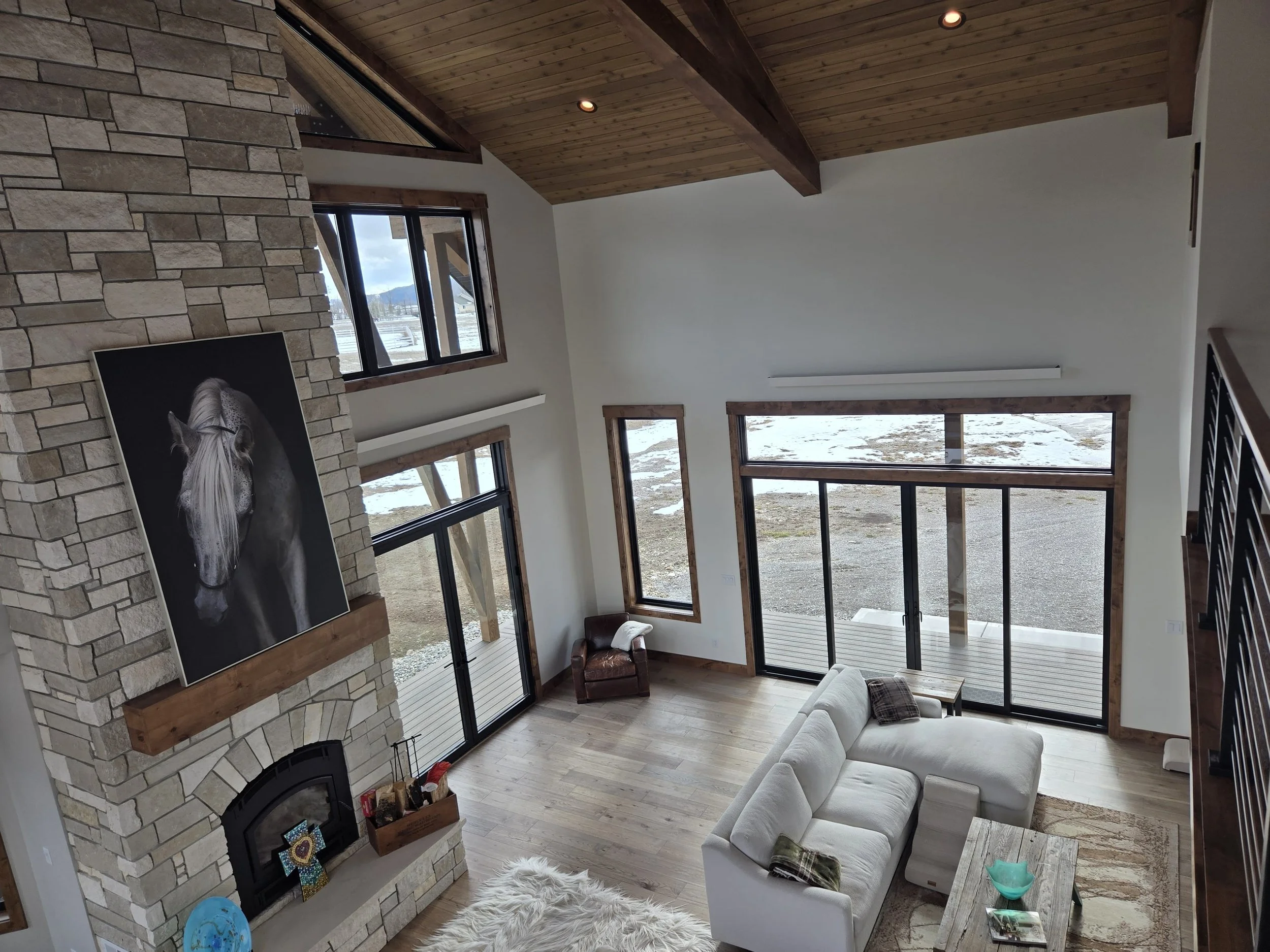 Living room with high vaulted wood ceiling and large windows, stone fireplace with a black and white horse portrait, beige sectional sofa, sliding glass doors leading to outdoor deck, and mountain landscape visible outside.