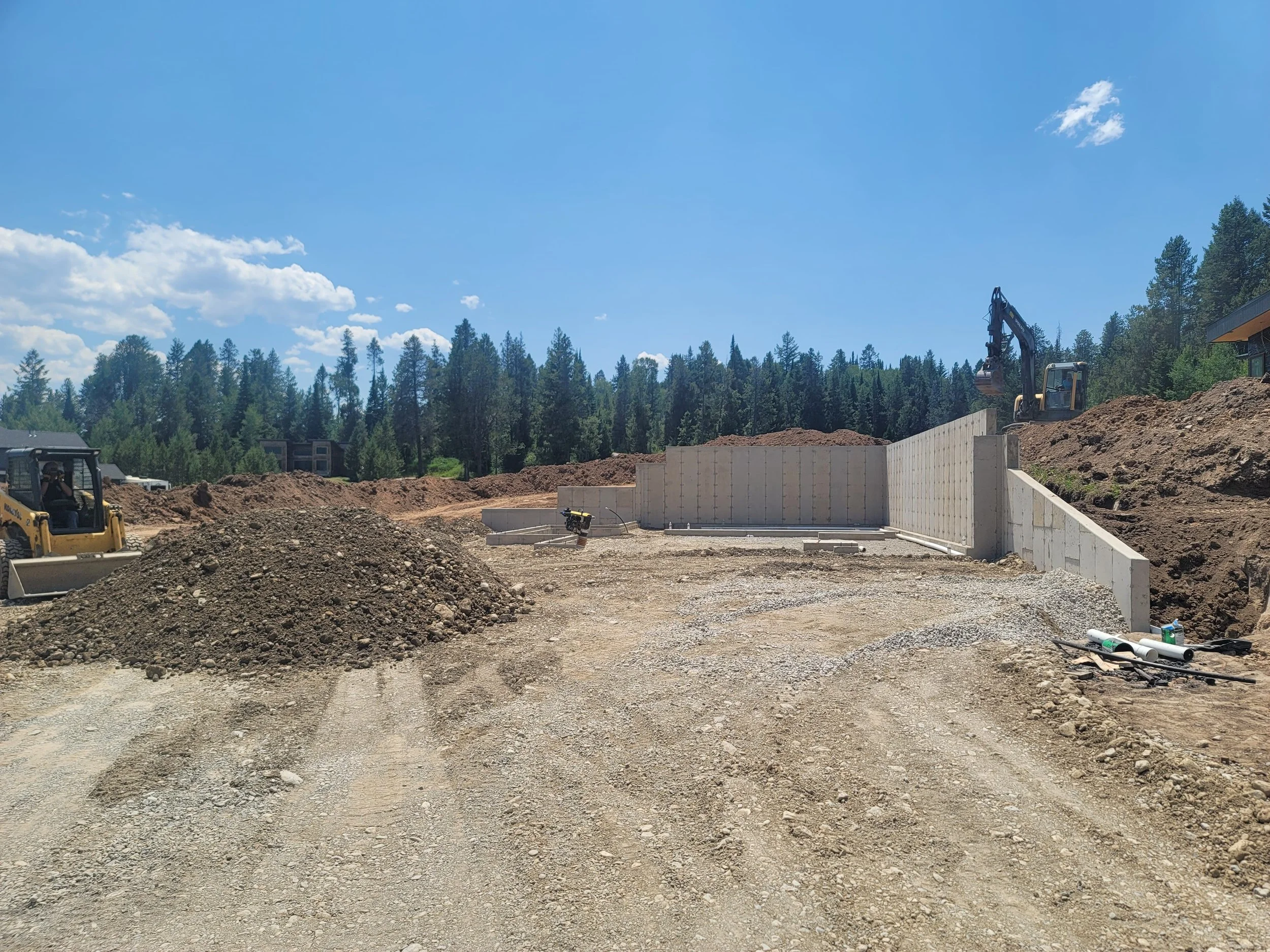 Construction site with dirt piles, a partially built concrete wall, and excavators on a sunny day with a blue sky and trees in the background.