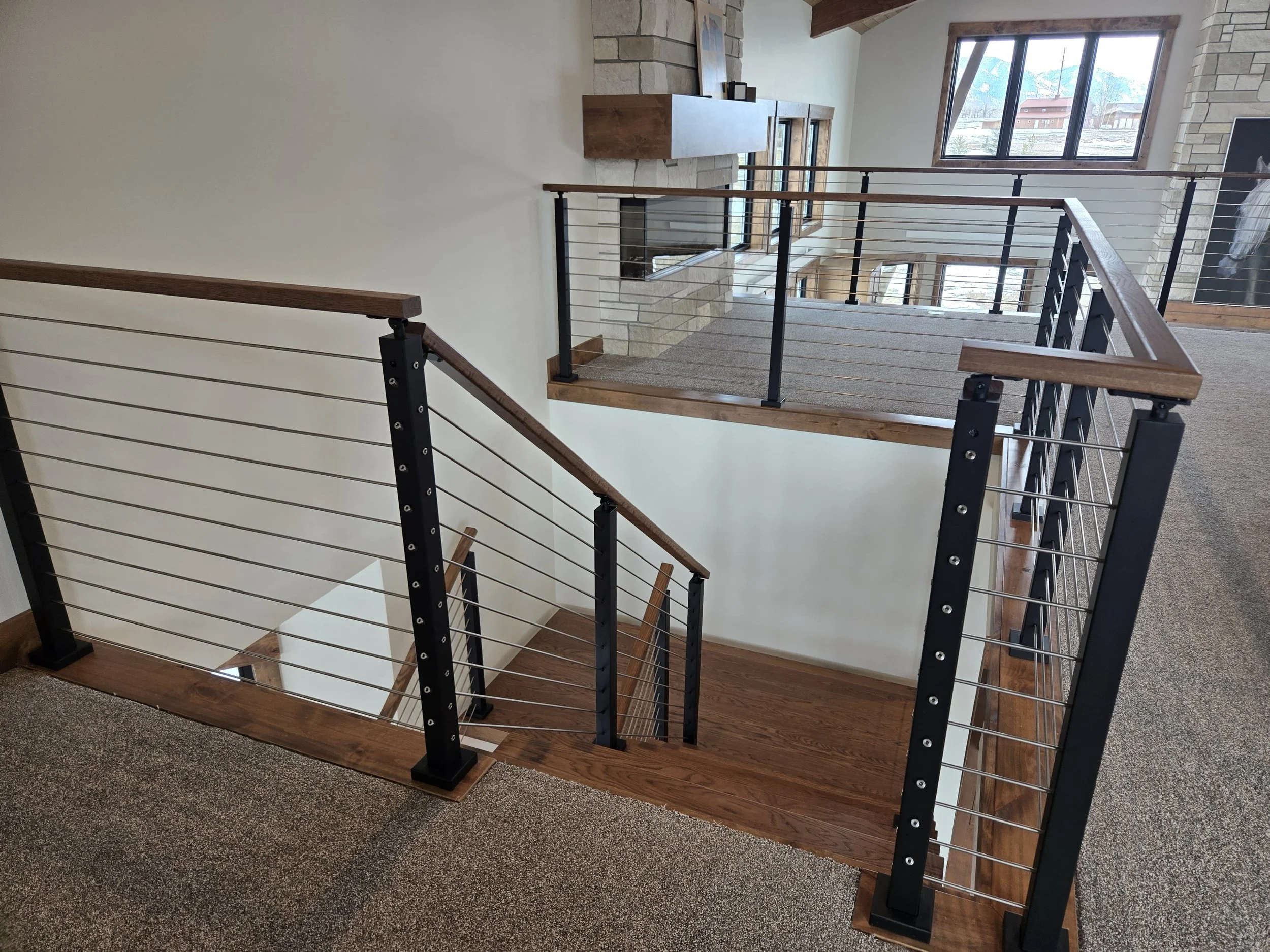 Interior view of a staircase with wooden handrails and metal balusters, overlooking a loft area with carpeted flooring, large windows, and stone walls, in a modern home.