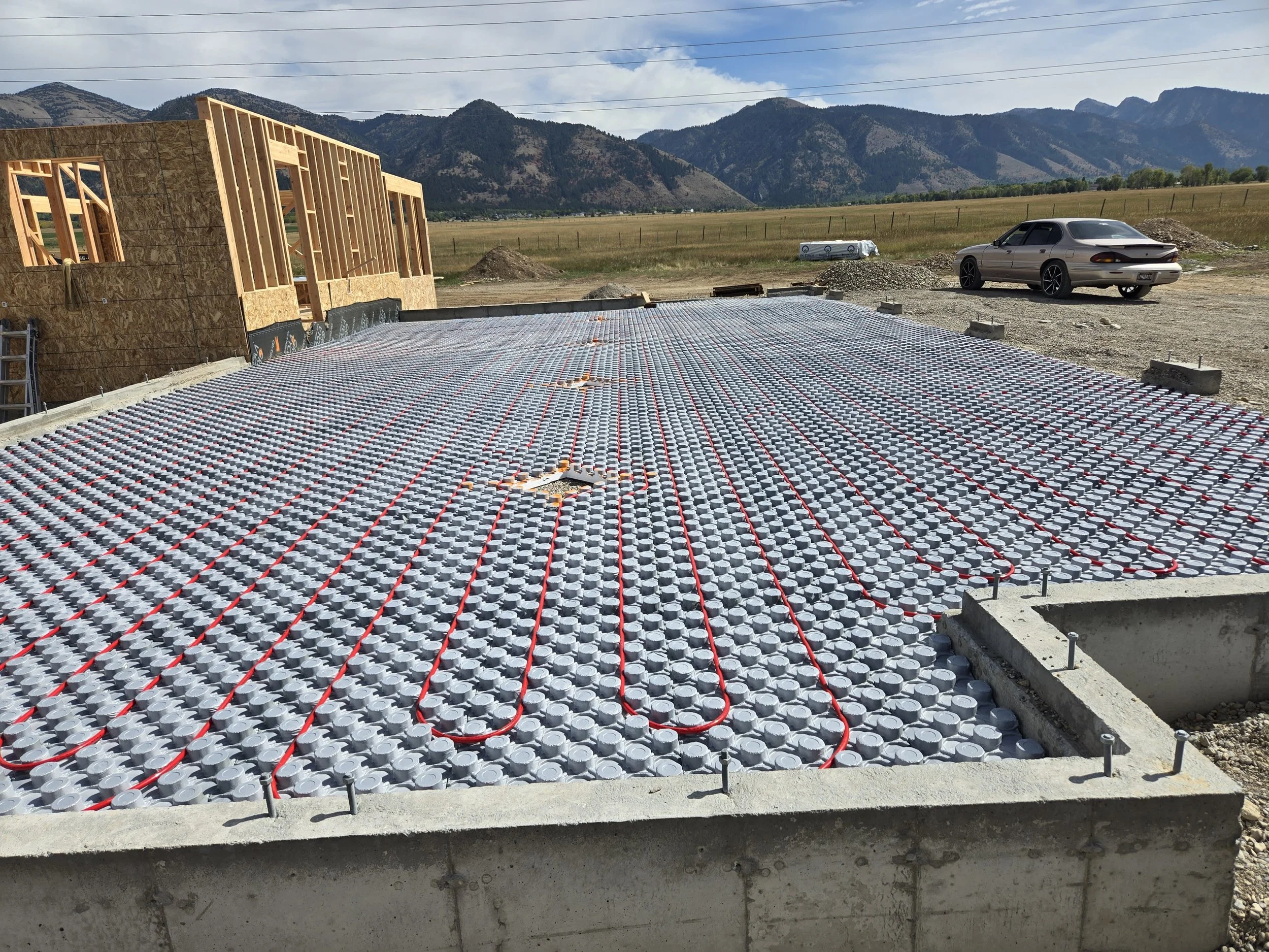 Construction site with a foundation ready for concrete, with underfloor radiant heating system installed, and a car parked nearby with mountains in the background.