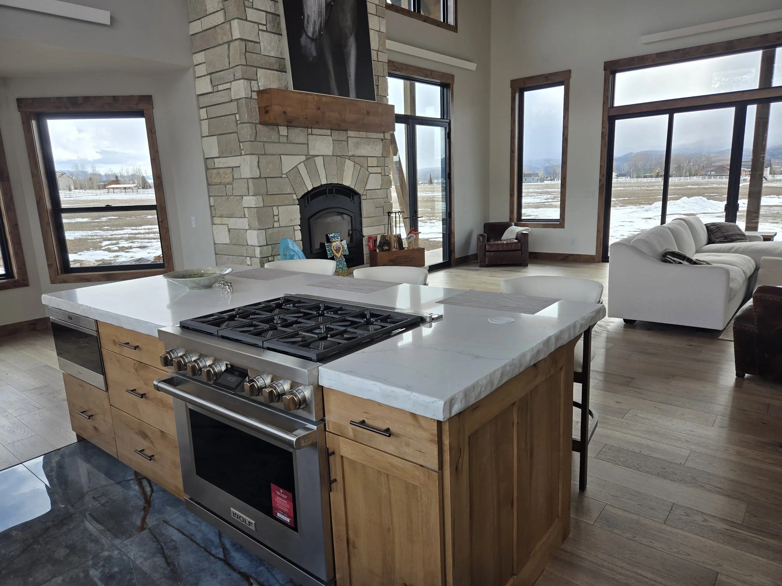 Modern open-concept kitchen and living room with large windows, stone fireplace, white sofas, and wooden floors, overlooking a snowy landscape in Thayne, WY.