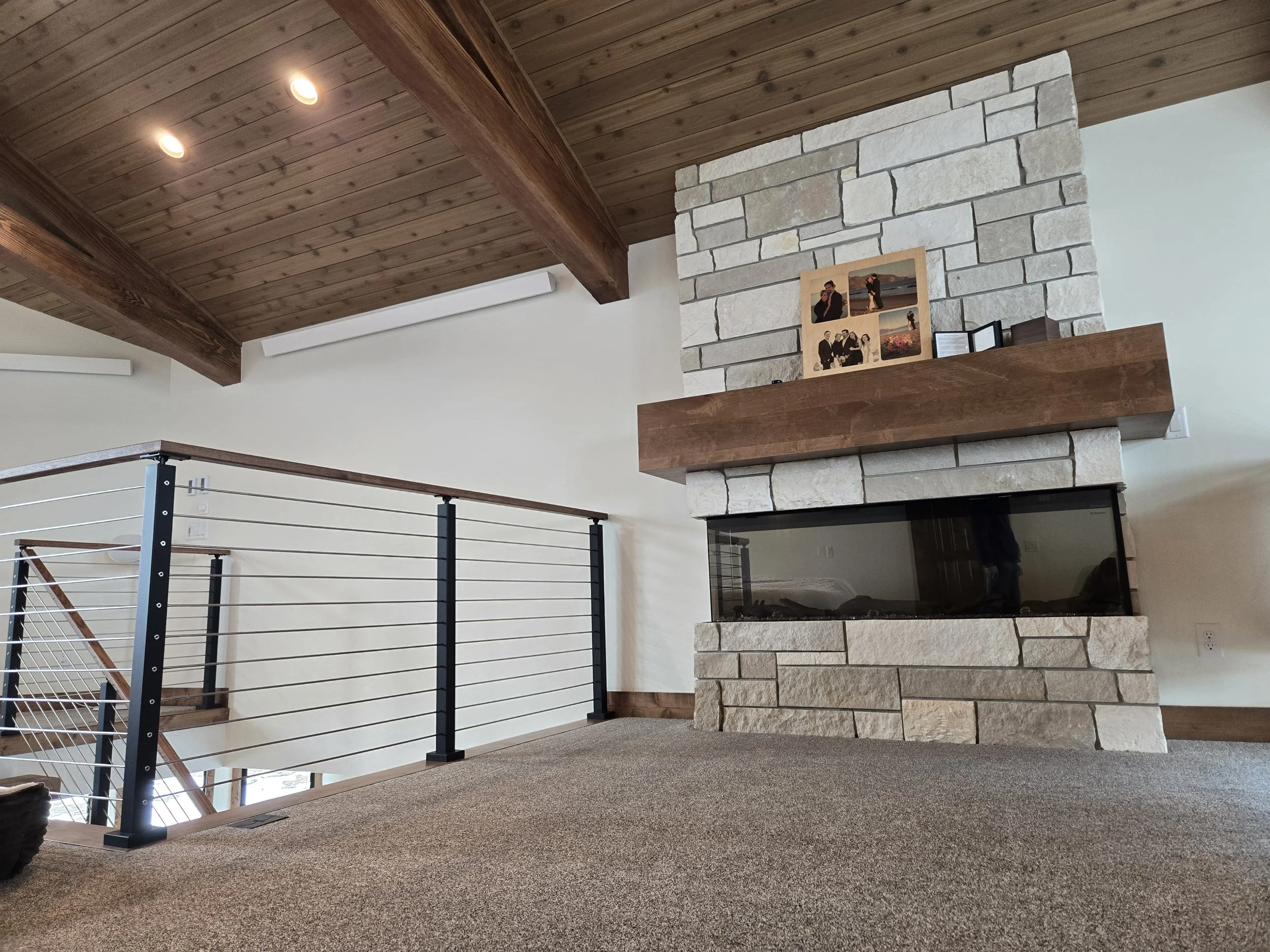 Interior of a house with a stone fireplace, wooden mantel, and a view of a staircase with a metal and wood railing. The ceiling is wooden with recessed lighting, and the floor is carpeted.
