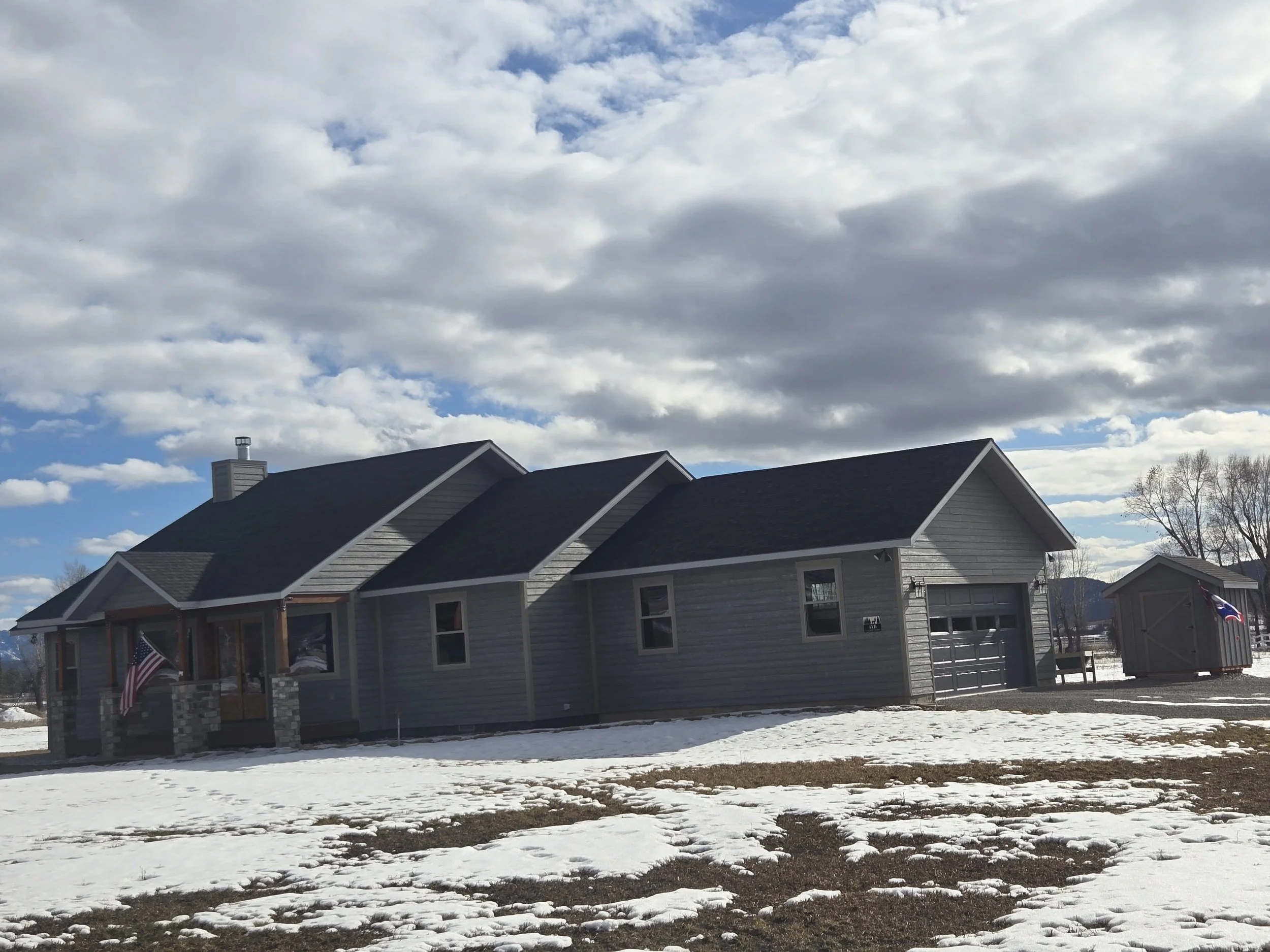 A gray house with a dark roof, a front porch with pillars, and a garage, set against a partly cloudy sky with snow on the ground and a small shed nearby.