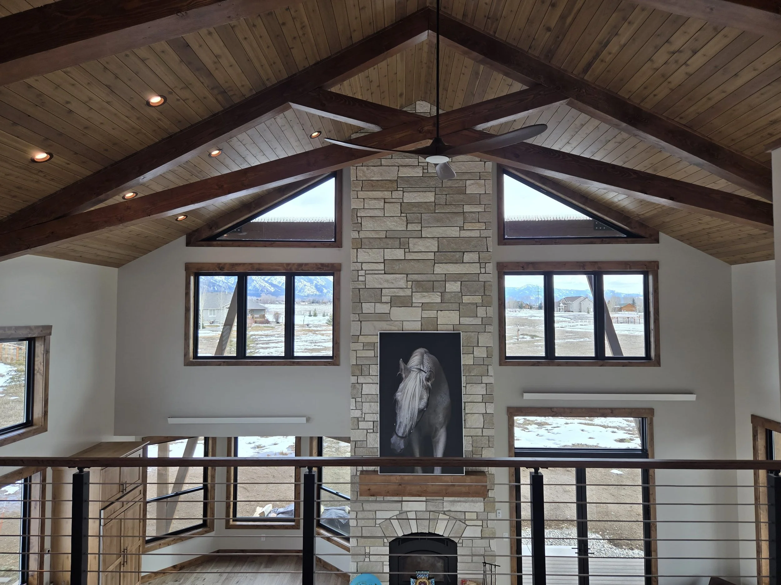 Interior view of a spacious living room with a vaulted wooden ceiling, a stone fireplace, a ceiling fan, and large windows showing a mountainous landscape outside.