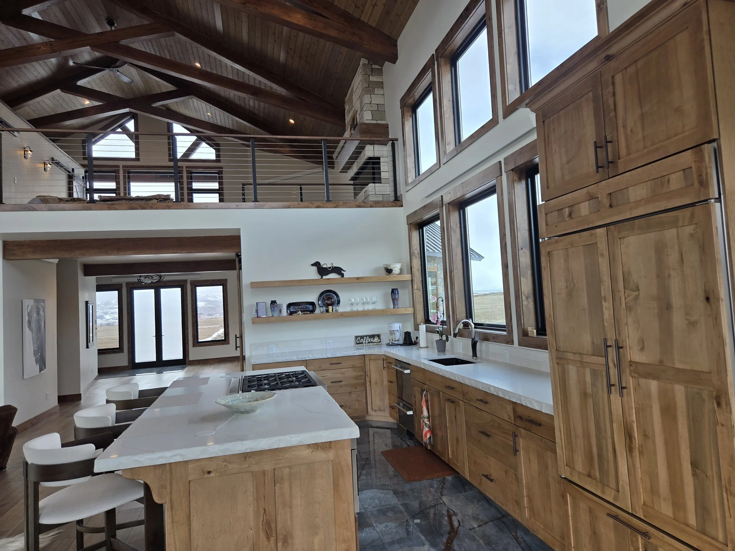 Kitchen with wooden cabinets, white countertops, large windows, and modern appliances. An island with white chairs is in the foreground.