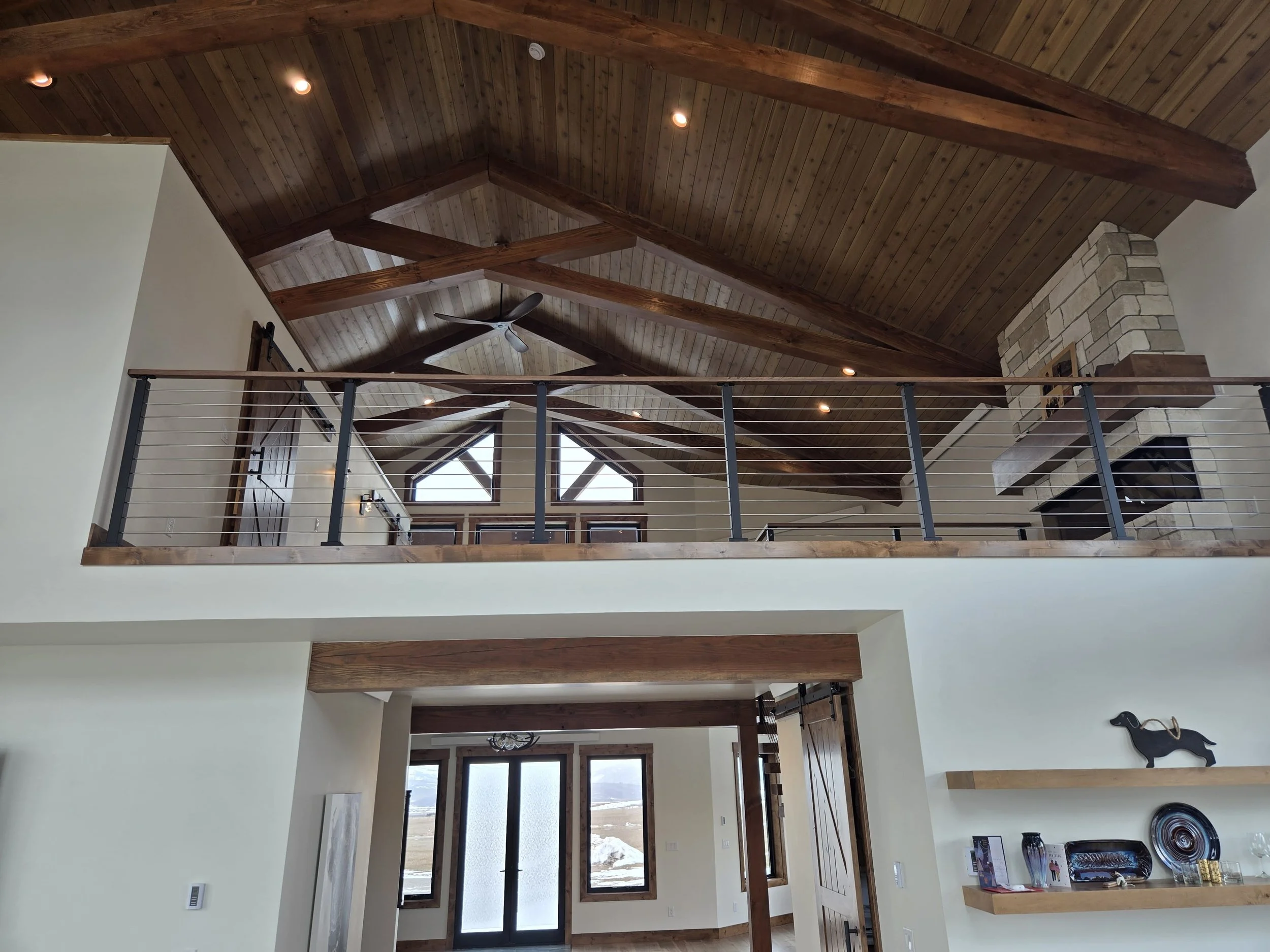 Interior view of a spacious living area with a vaulted wooden ceiling, exposed wooden beams, a ceiling fan, a stone fireplace, large windows, and a balcony with metal railing.