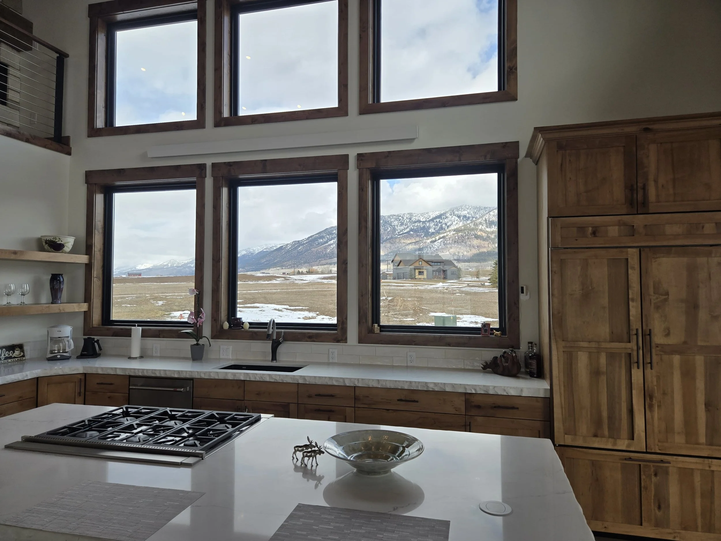 Kitchen with large windows overlooking snowy mountains and open field, wooden cabinetry, white island countertop, and decorative items.