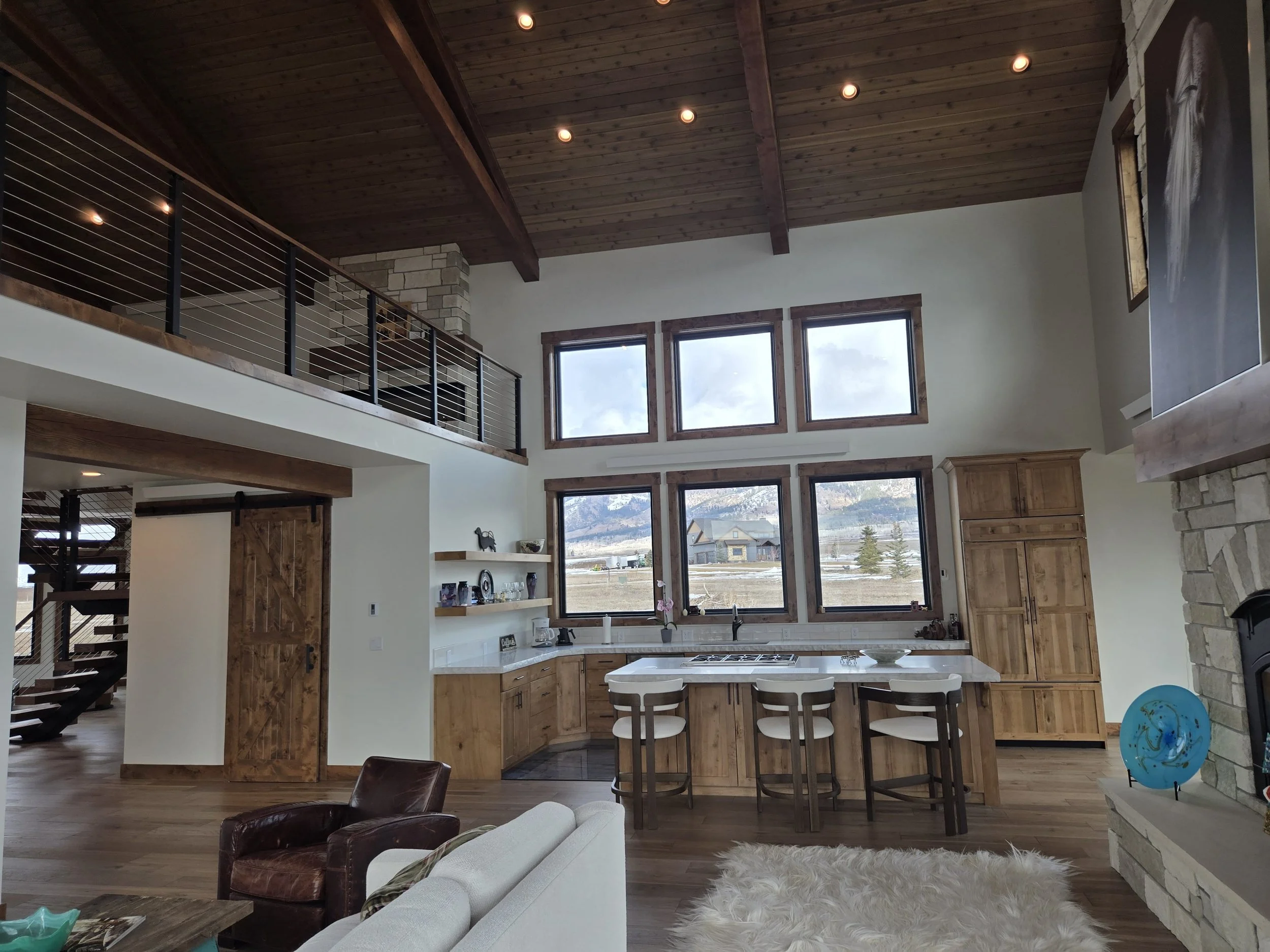 Open-concept living space with high wooden ceiling, large windows, and a kitchen island with stools, a leather armchair, a white sofa, a stone fireplace with a blue decorative plate, and built-in wooden cabinets.