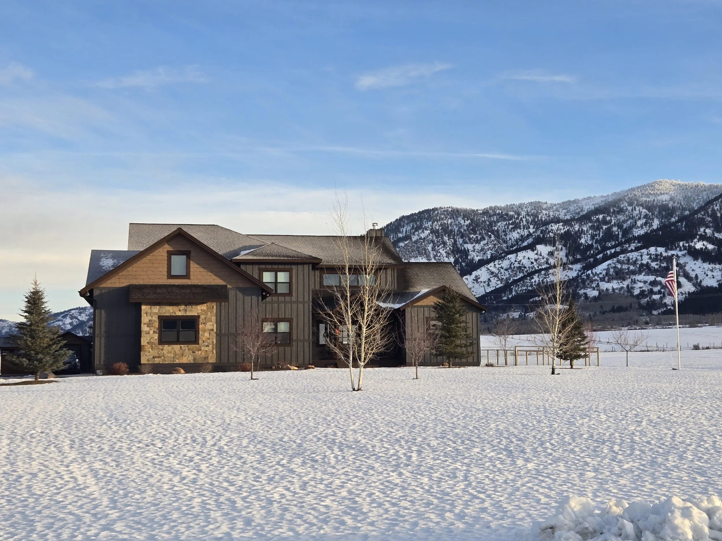 A house with a stone and wood exterior surrounded by snow, with mountains in the background and a flagpole with the American flag.