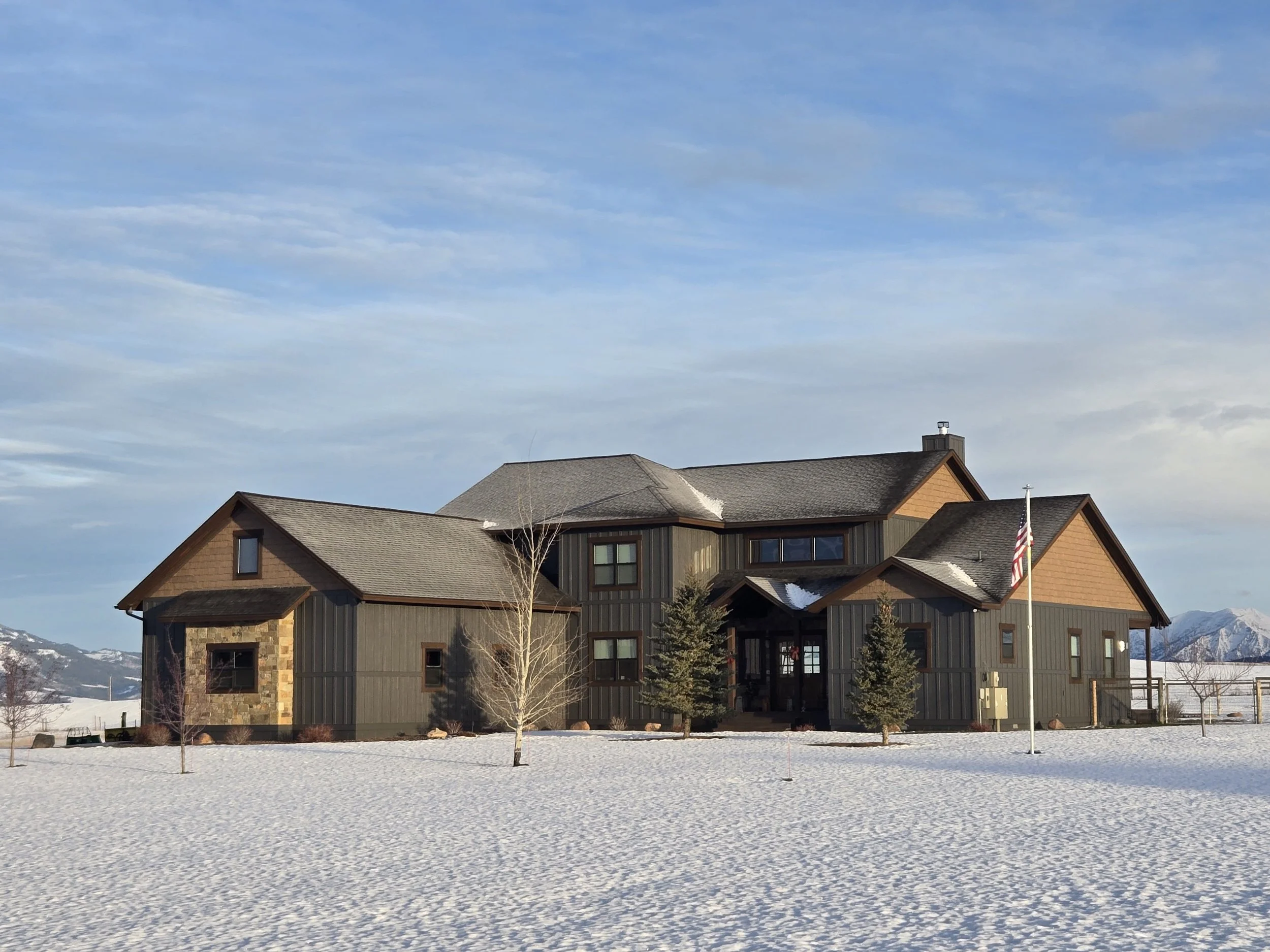 A large house with dark wooden siding and a stone foundation, surrounded by snow, with a flagpole, several trees, and mountains in the background under a cloudy sky.
